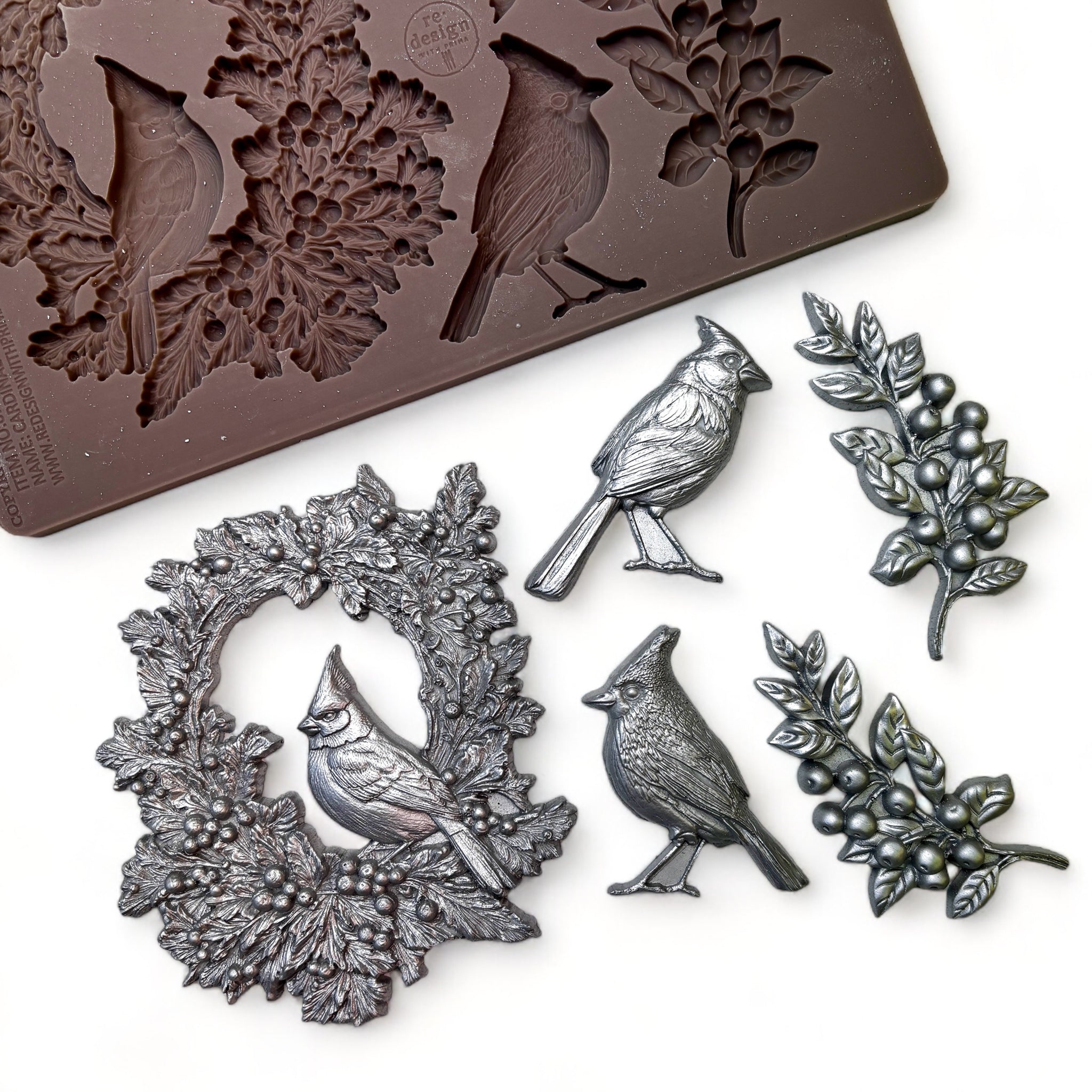 A brown silicone mold and silver colored castings of a festive wreath with a resting cardinal, as well as two individual cardinals and two sprigs of greenery are against a white background.