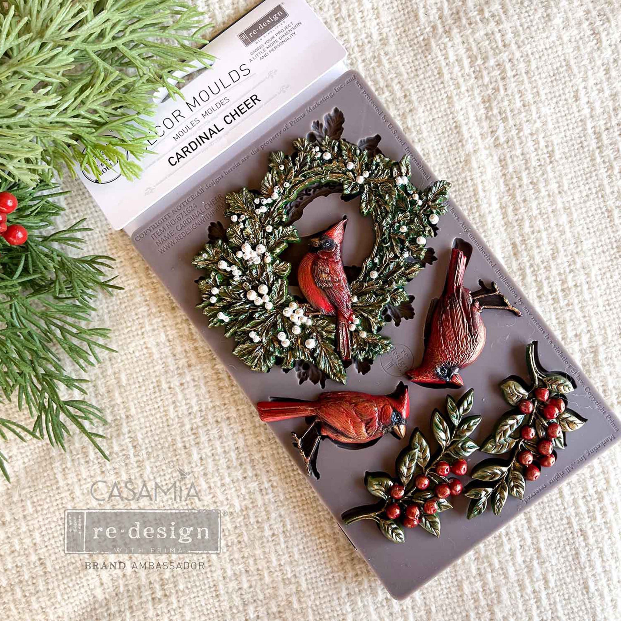 A package of ReDesign with Prima's Festive Cardinal Cheer silicone mold and colorful castings created by Casamia are against a white linen background.