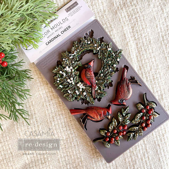 A package of ReDesign with Prima's Festive Cardinal Cheer silicone mold and colorful castings created by Casamia are against a white linen background.