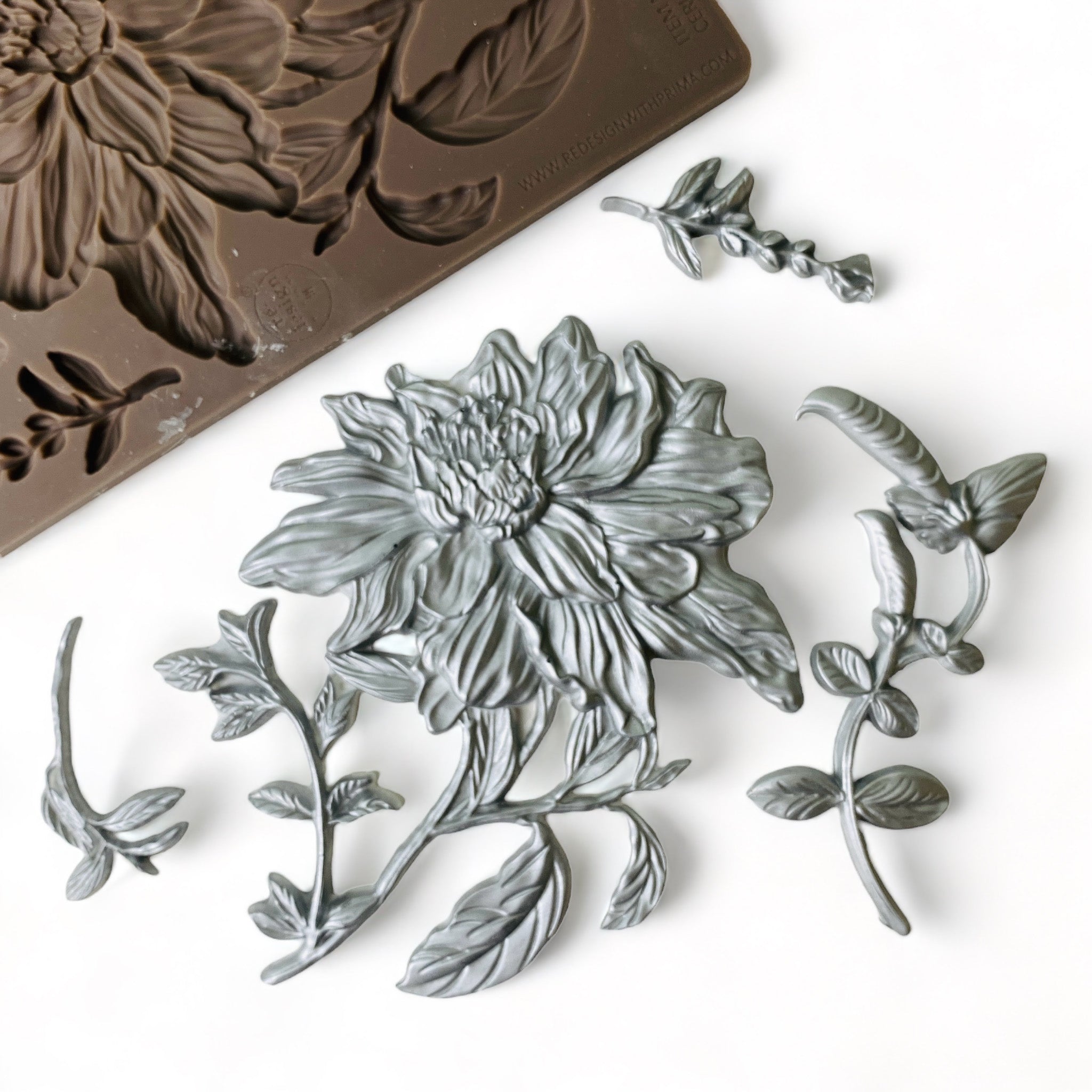 A brown silicone mold and silver-colored castings of a large flower bloom with foliage are against a white background.