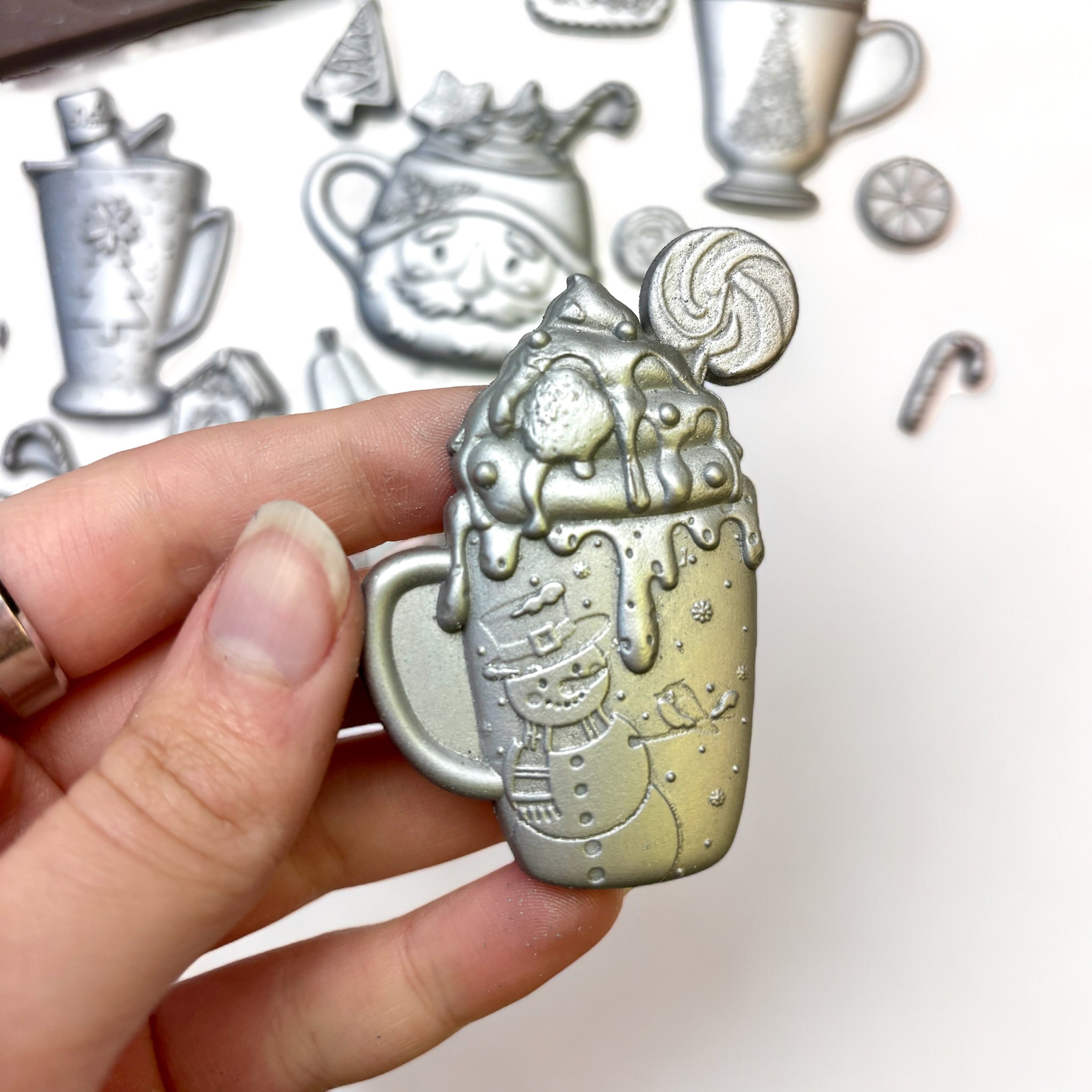 Close-up of a hand holding a silver colored casting from ReDesign with Prima's Cheerful Cups silicone mold is shown against a white background.