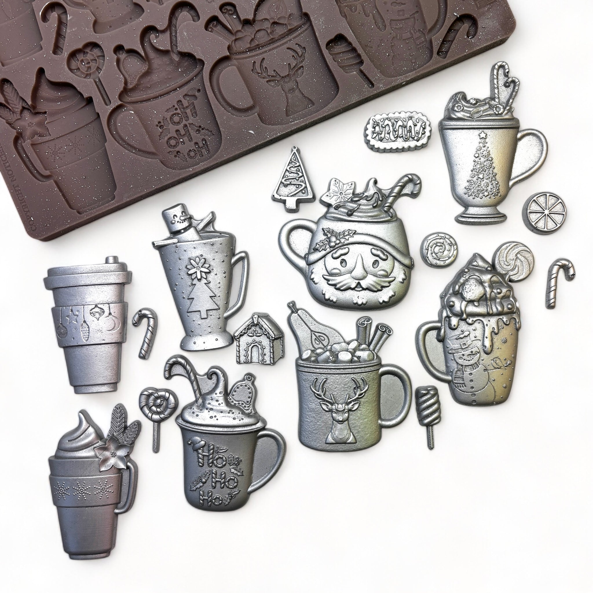 A brown silicone mold and silver colored castings of 8 mugs of favorite holiday drinks and 9 adorable shapes like candy canes and gingerbread houses are against a white background.