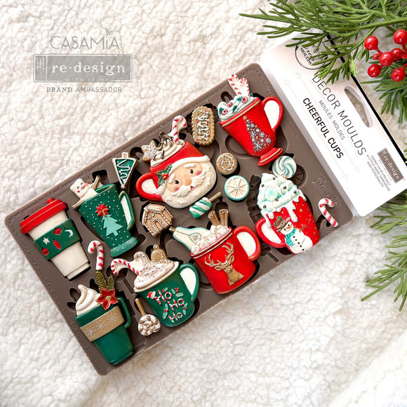 A package of ReDesign with Prima's Cheerful Cups silicone mold and colorful castings made by Casamia are against a white Sherpa blanket.