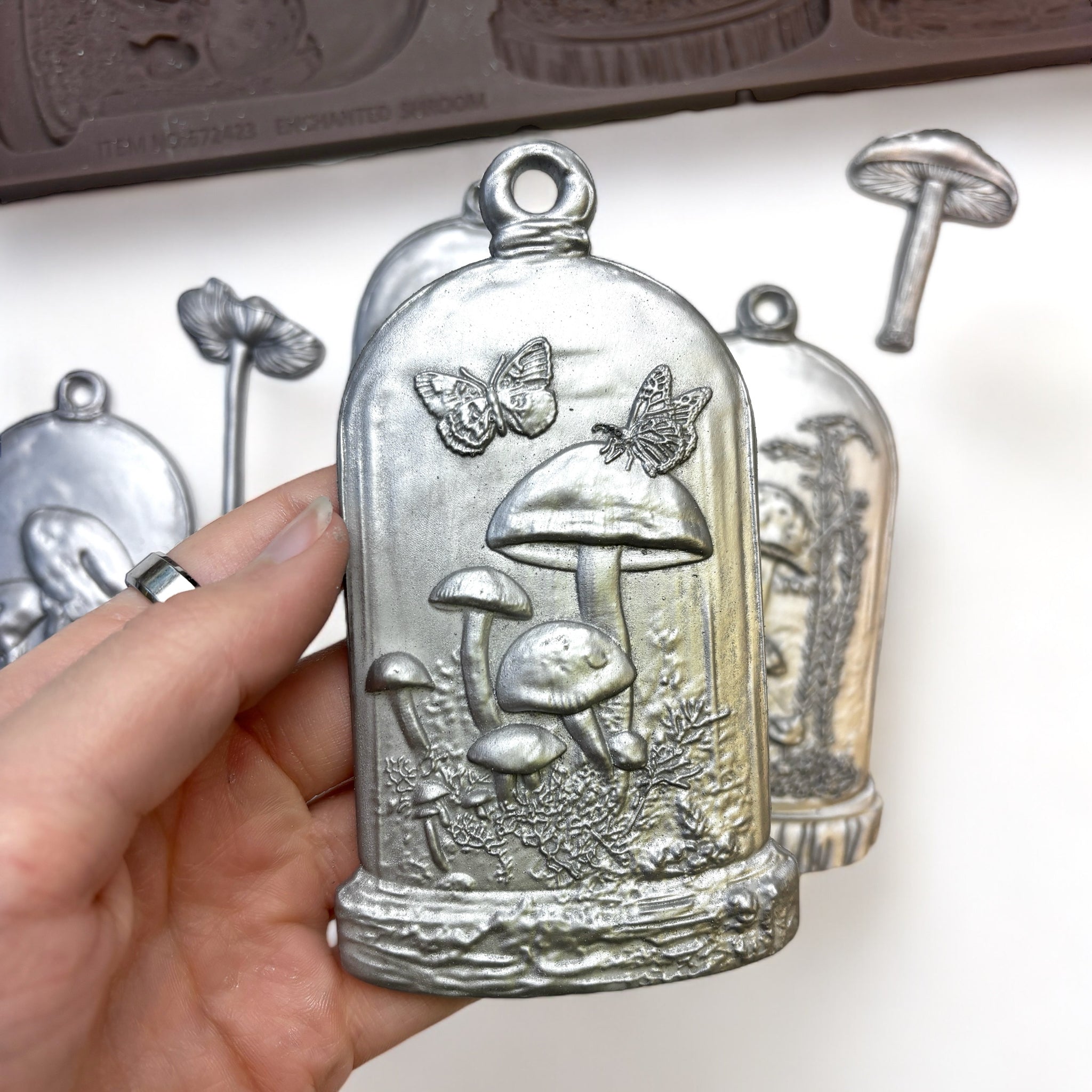 A close-up of a hand holding a silver colored casting from ReDesign with Prima's Enchanted Shroom silicone mold is shown against a white background.