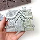 A close-up of a hand shown holding a silver colored casting of a gingerbread house created from ReDesign with Prima's Festive Gingerbread Treats silicone mold is against a white background.