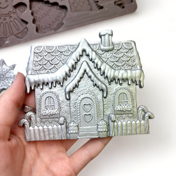 A close-up of a hand shown holding a silver colored casting of a gingerbread house created from ReDesign with Prima's Festive Gingerbread Treats silicone mold is against a white background.