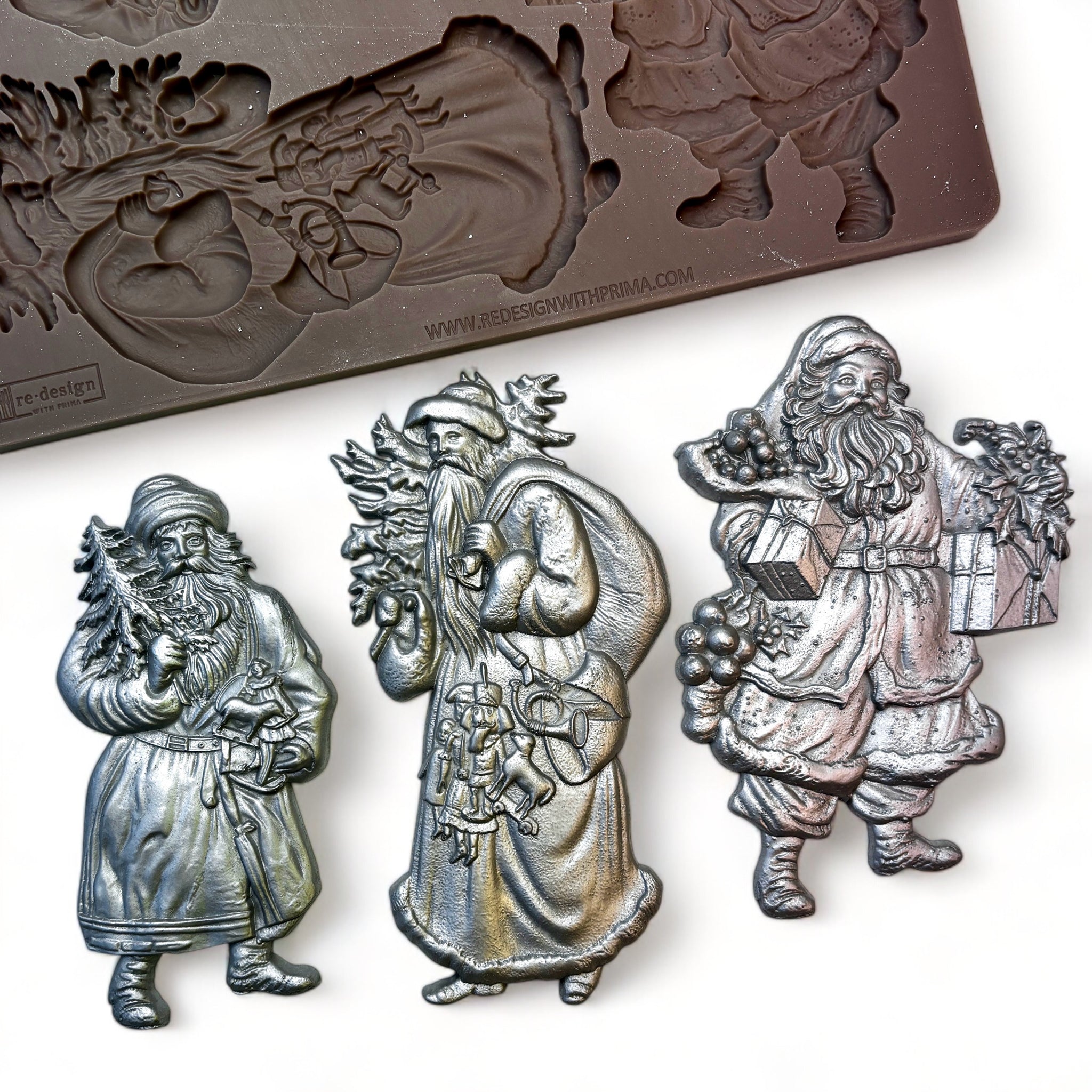 A brown silicone mold and silver colored castings of 3 festive designs of Santa Claus with Christmas gifts and greenery are against a white background.