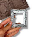 Hand holding a small ornate silver mirror with a silicone mold in the background