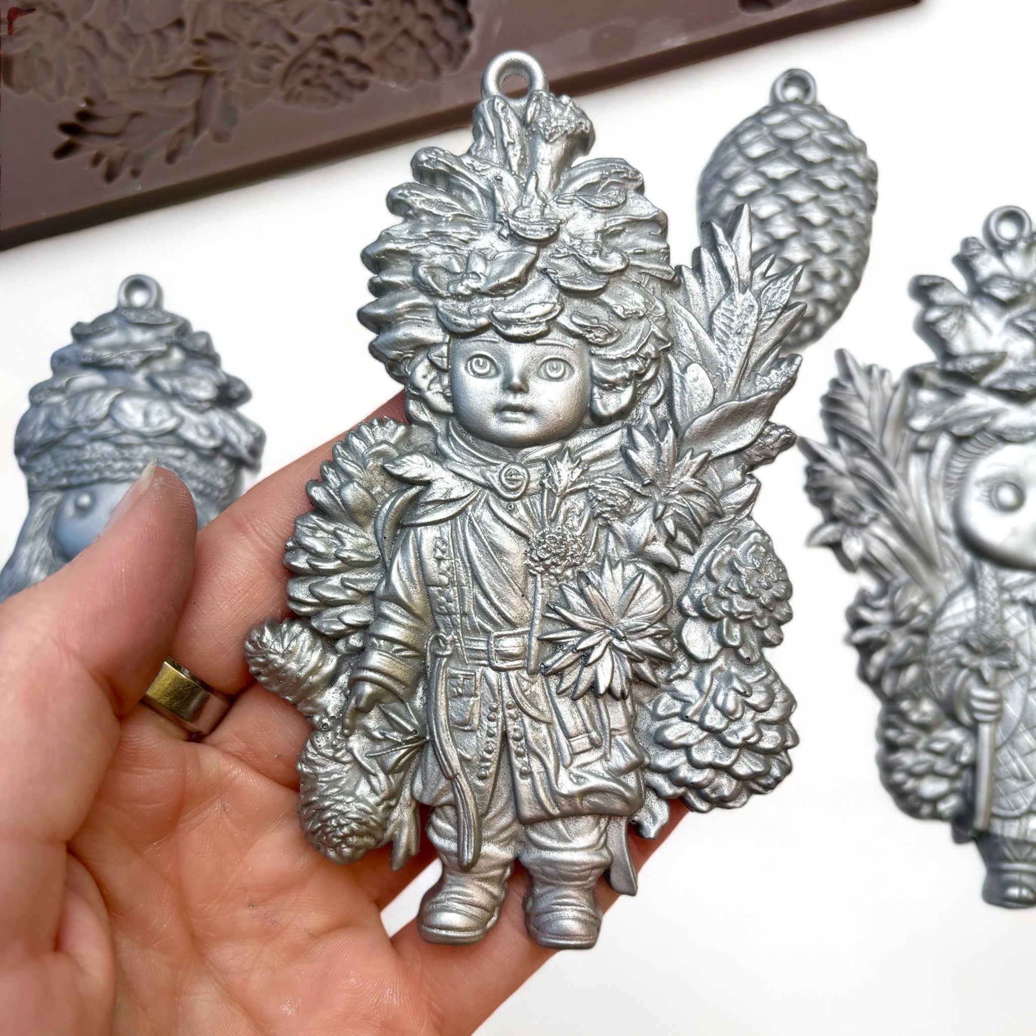 A close-up of a hand holding a silver colored casting of a pinecone doll made from ReDesign with Prima's Pinecone Dolls silicone mold is against a white background.