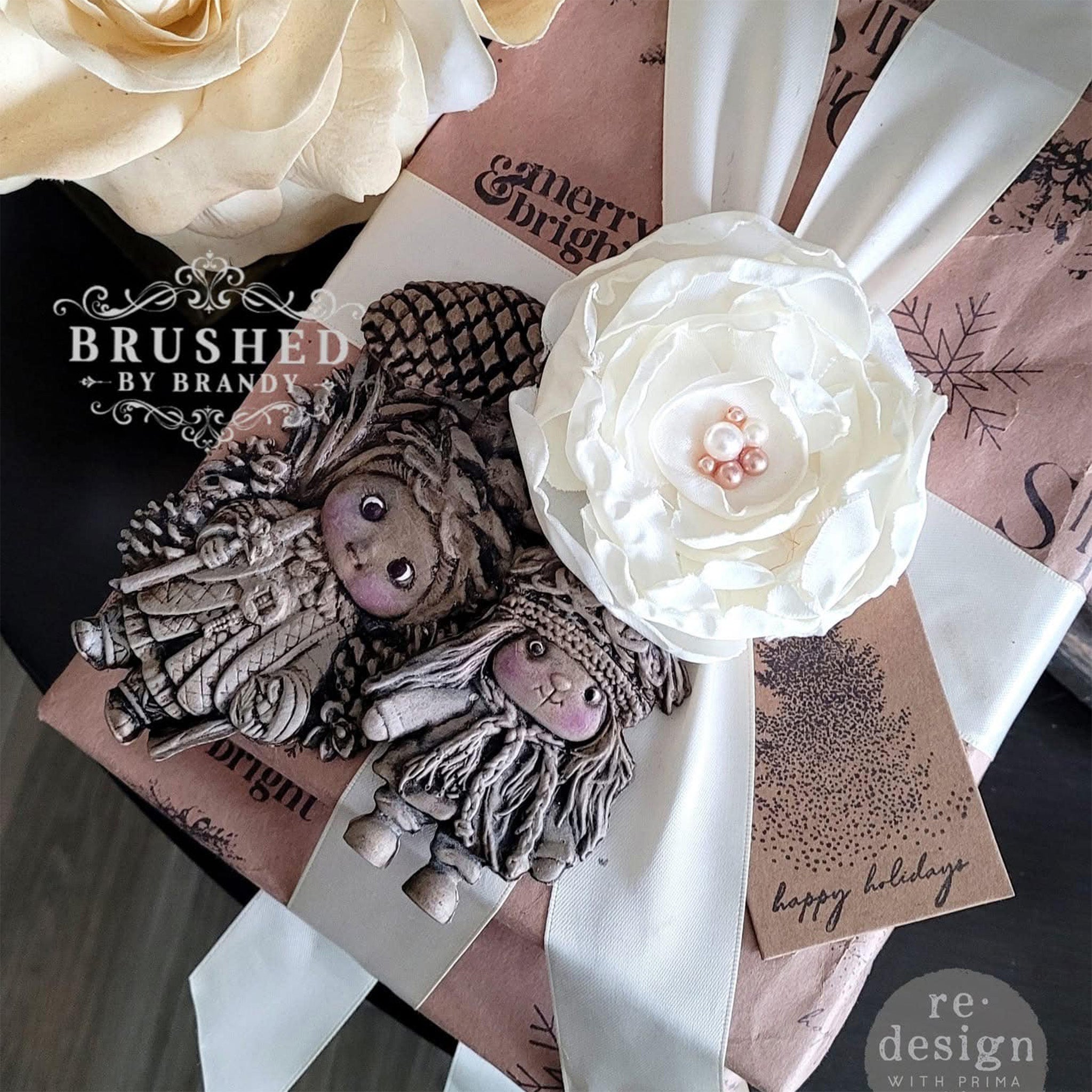 A wrapped gift with white ribbon and a silk white flower features castings of ReDesign with Prima's Pinecone Dolls silicone mold on it.