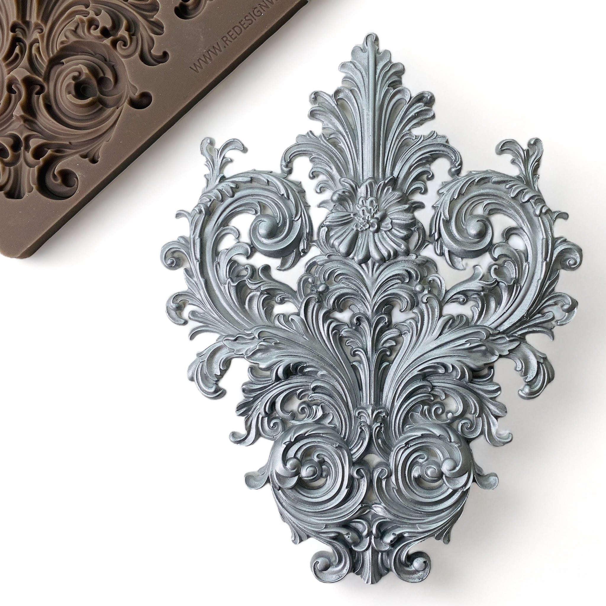 A brown silicone mold and silver-colored casting of an ornate leaf scroll design are against a white background.