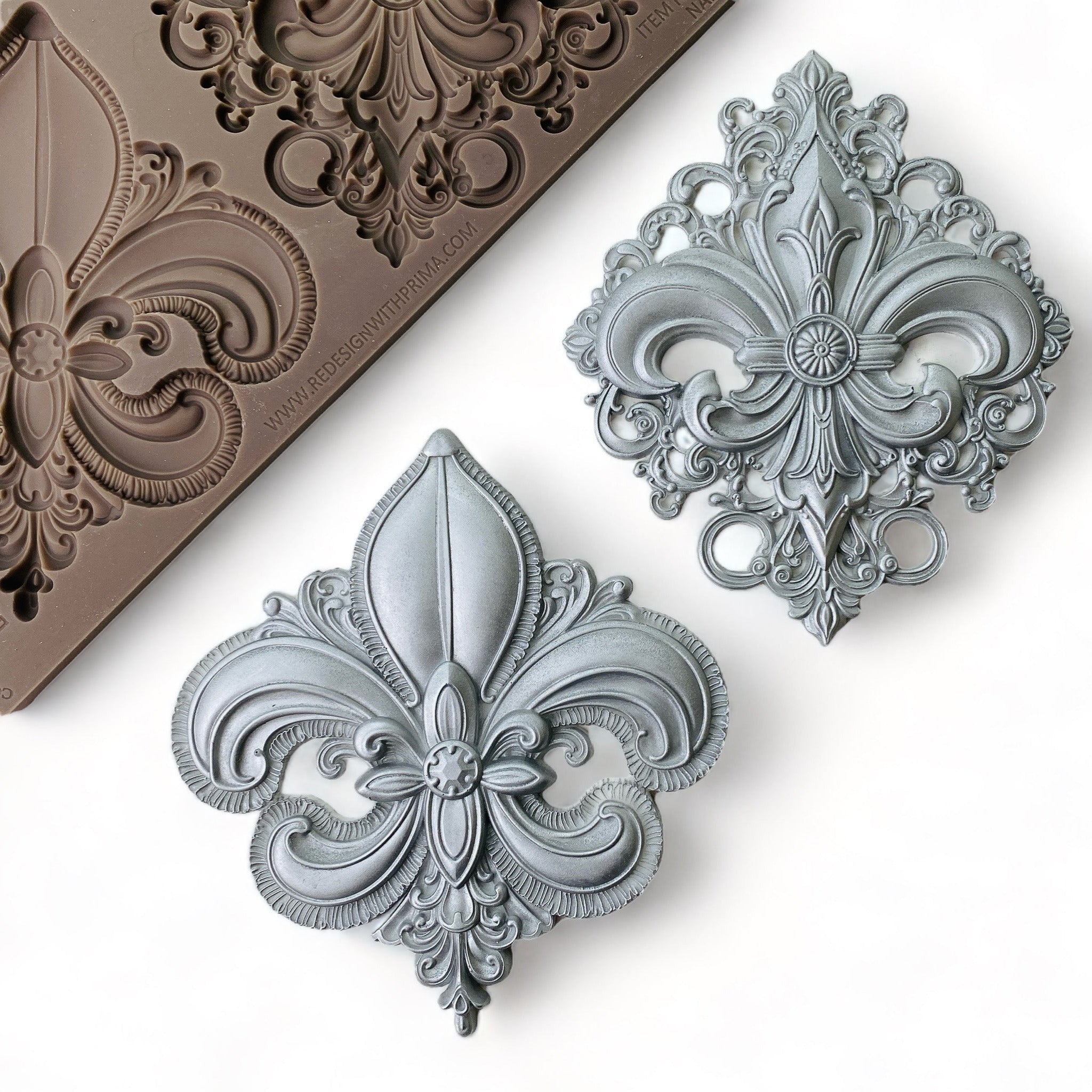 A brown silicone mold and silver-colored castings of 2 large ornate Fleur de Lis designs are against a white background.