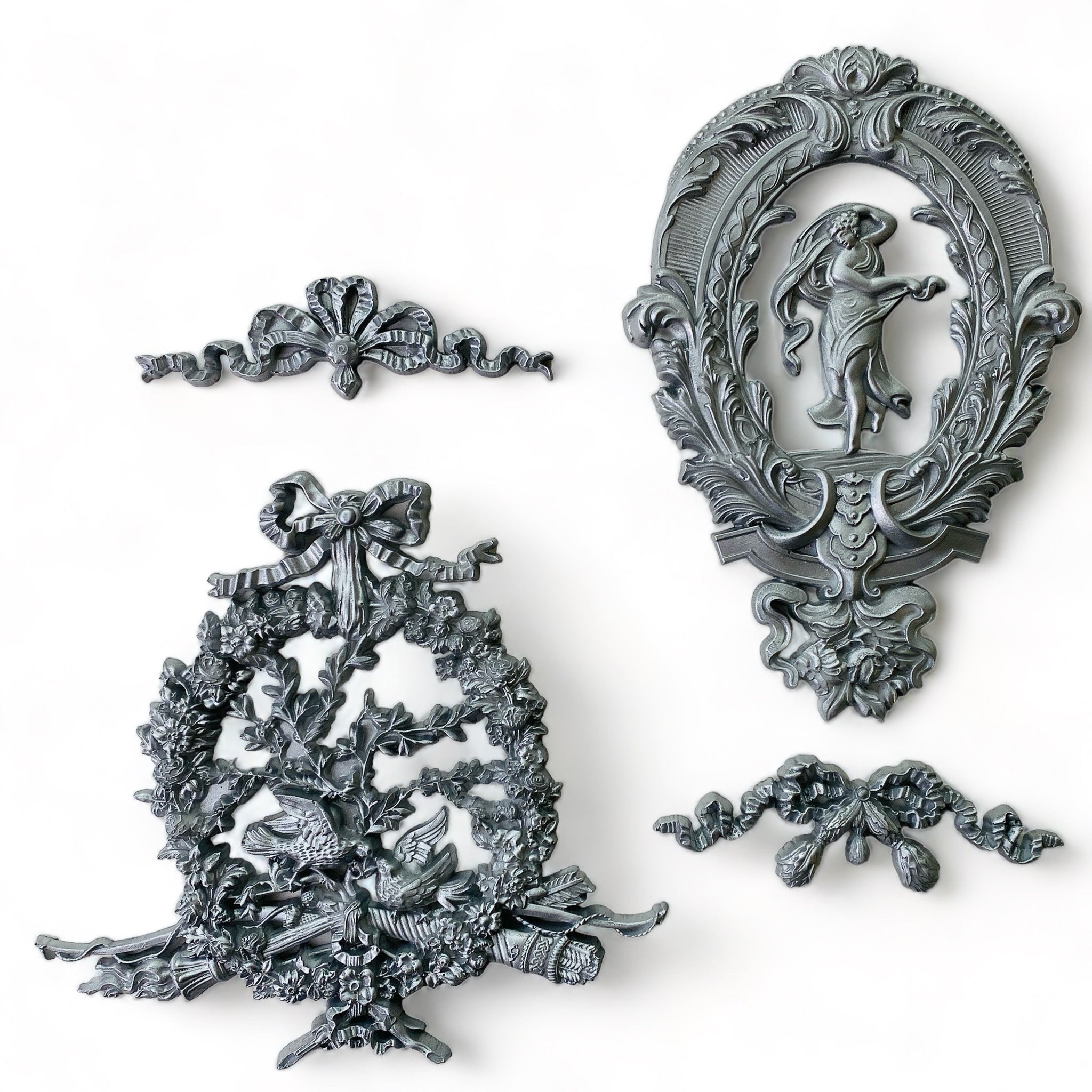 Silver colored silicone mold castings that feature 2 bows, an ornate oval medallion and an ornate wreath are against a white background.