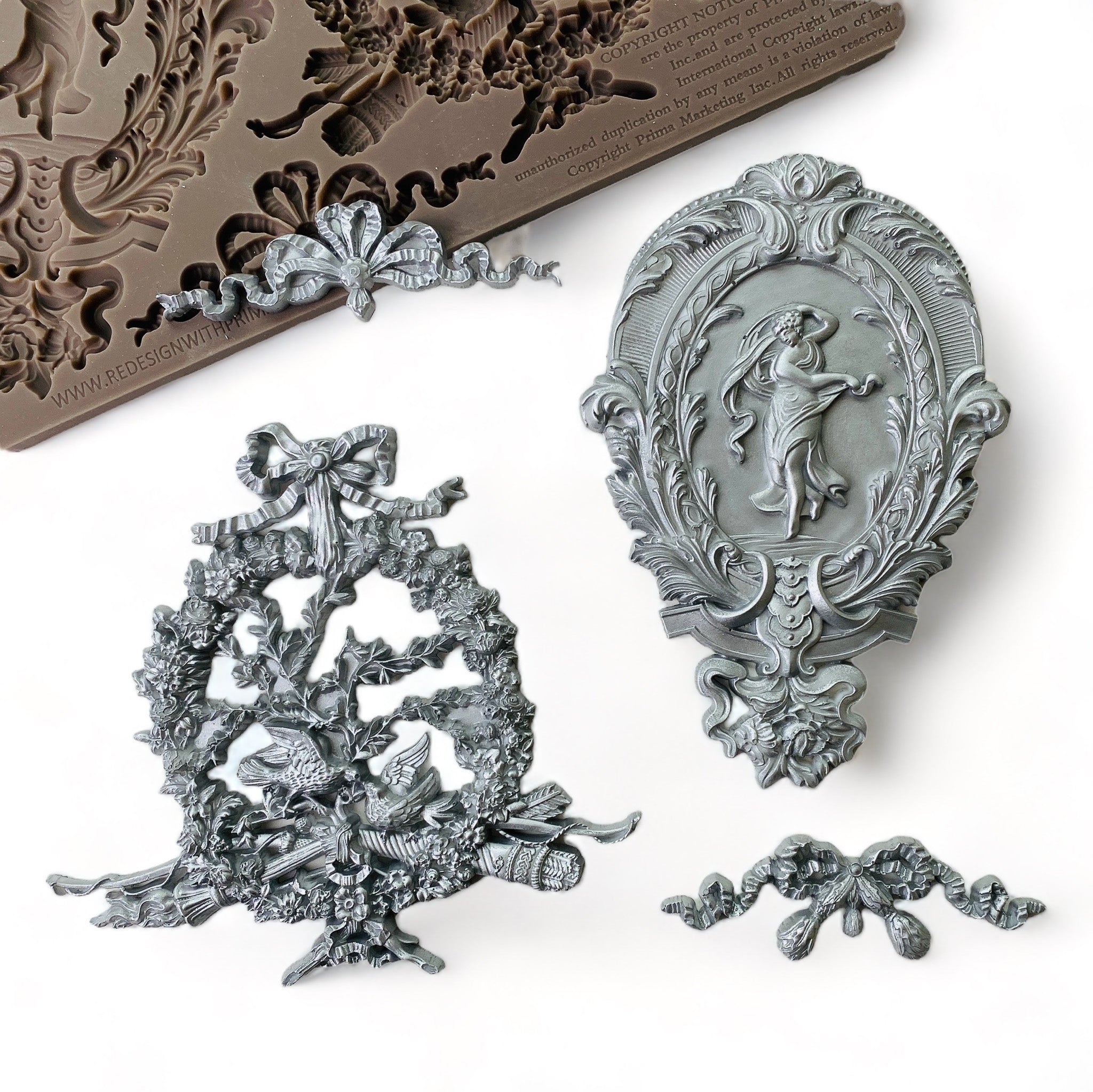 A brown silicone mold and its silver colored castings that feature 2 bows, an ornate oval medallion and an ornate wreath are against a white background.