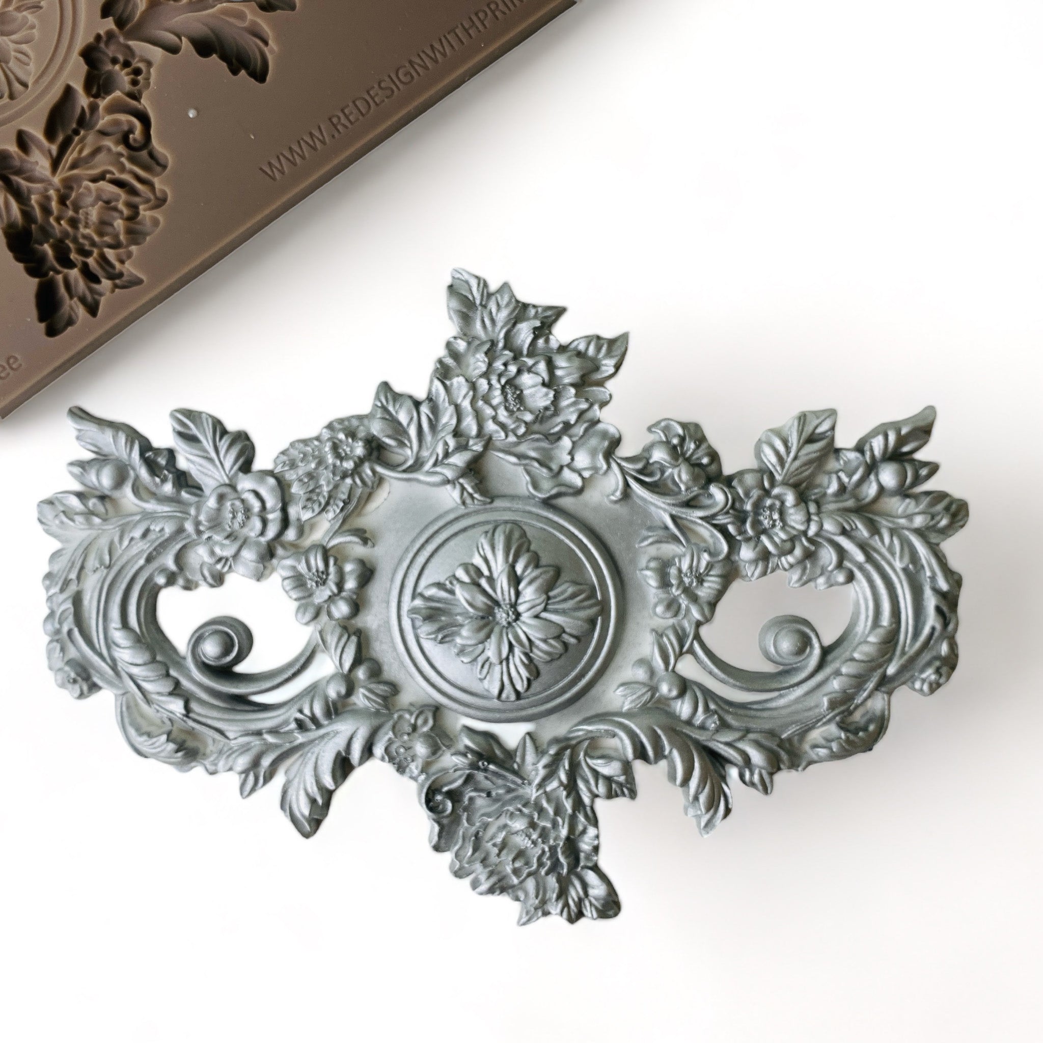 A brown silicone mold and silver-colored casting of a large ornate floral accented medallion center piece are against a white background.