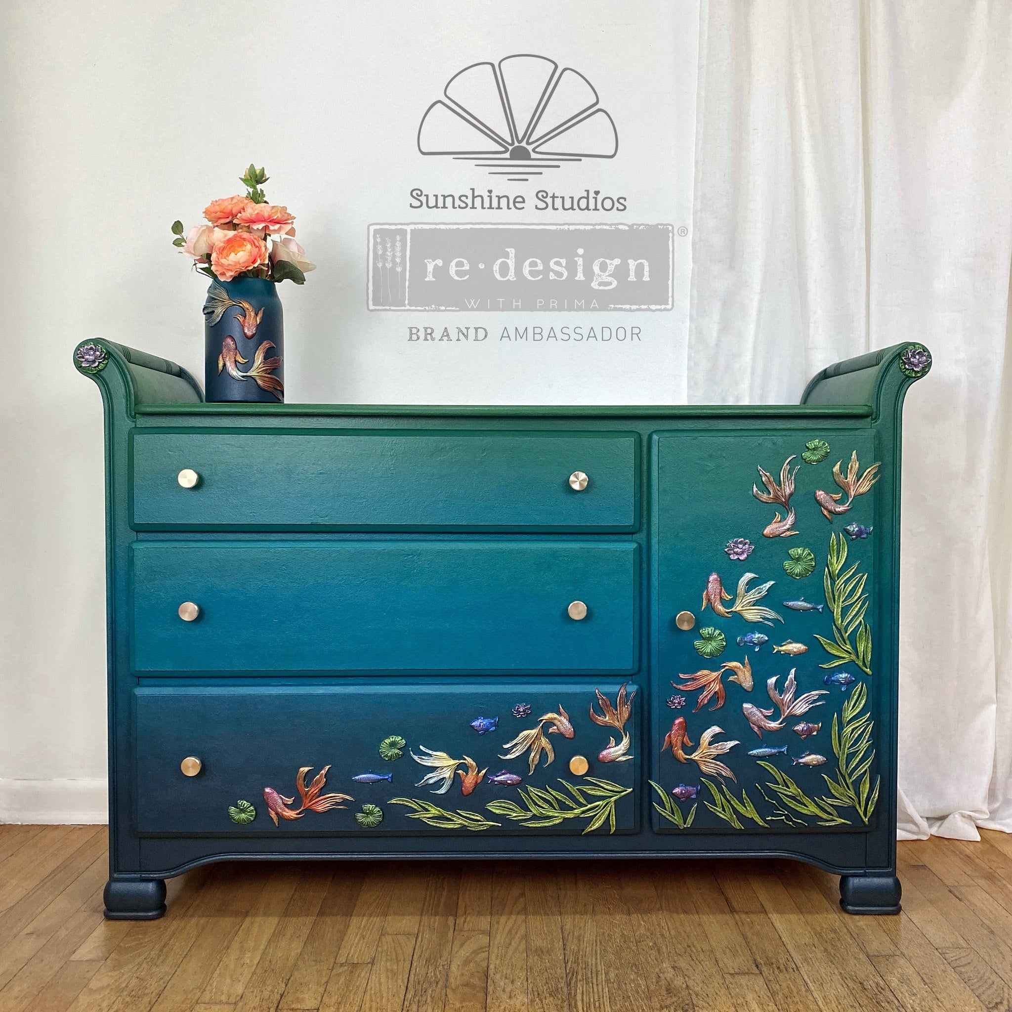 A vintage dresser refurbished by Sunshine Studios is painted a blend of teal and dark blue and features colorful castings from ReDesign with Prima's Siamese Splendor silicone mold on a door and drawer.