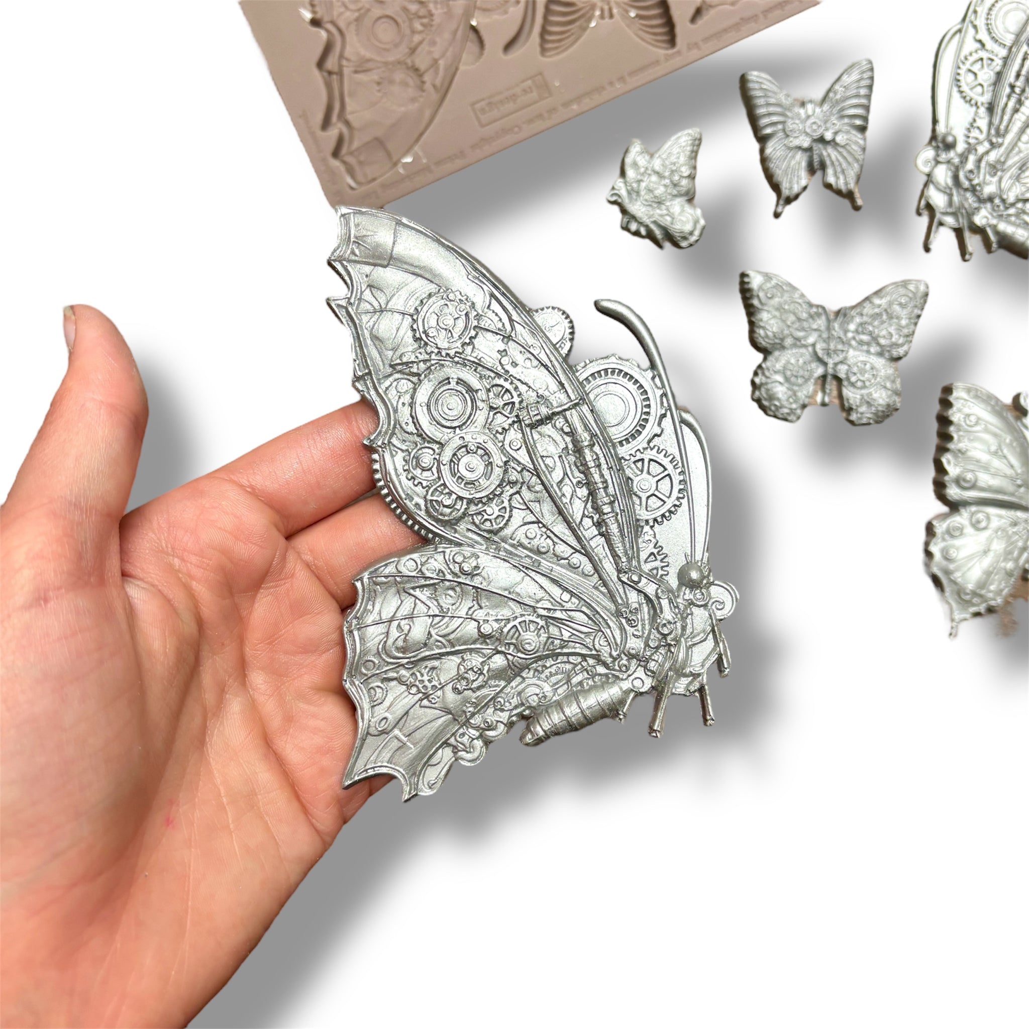 A silicone mold and silver colored castings featuring 6 intricate butterflies, each embellished with cogs and gears are against a white background. A hand is shown holding the largest casting.