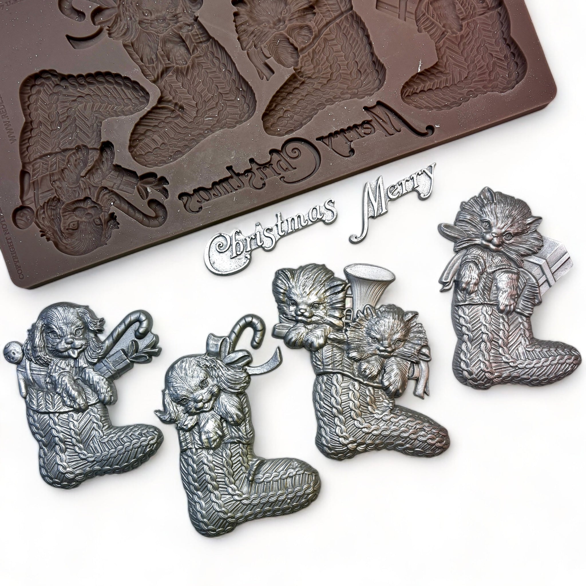 A brown silicone mold and silver colored castings of 4 Christmas stockings with puppies and kittens in them and the words Merry Christmas are against a white background.