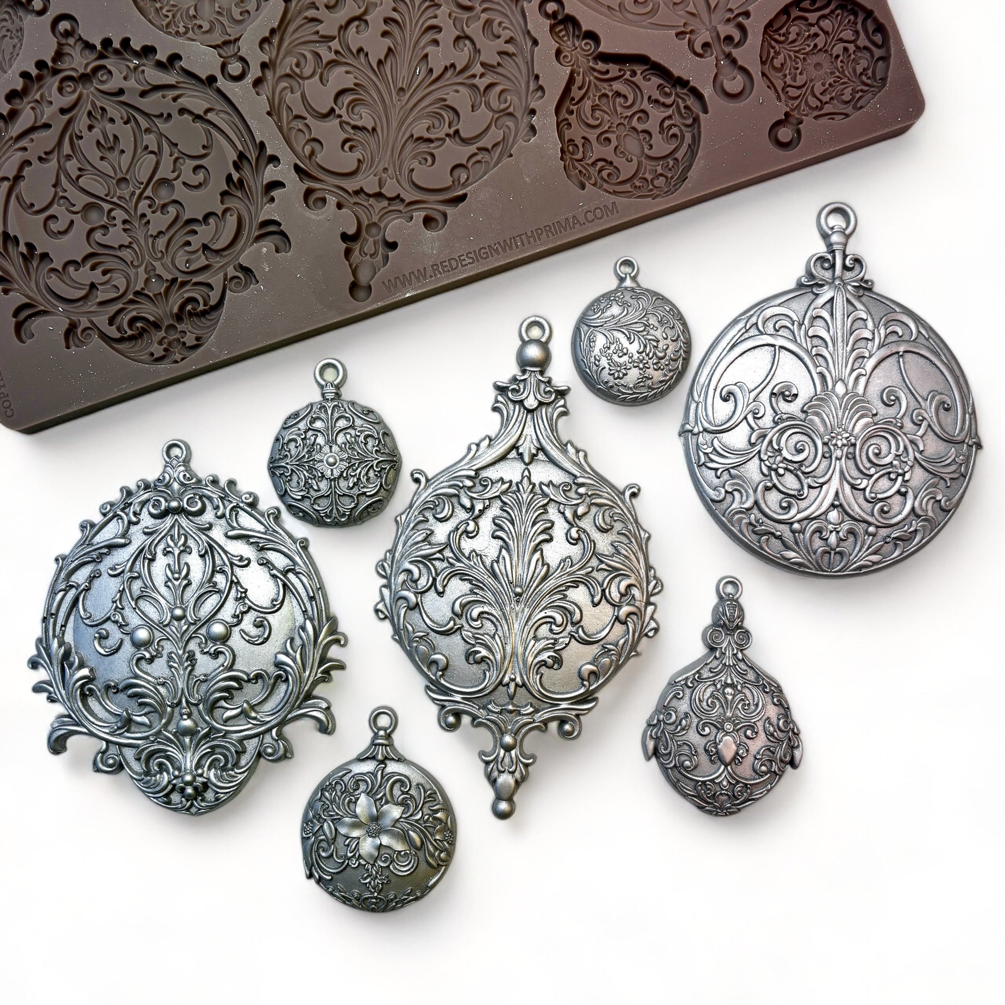 A brown silicone mold and silver colored castings of 3 large, and 4 small ornate ornaments are against a white background.