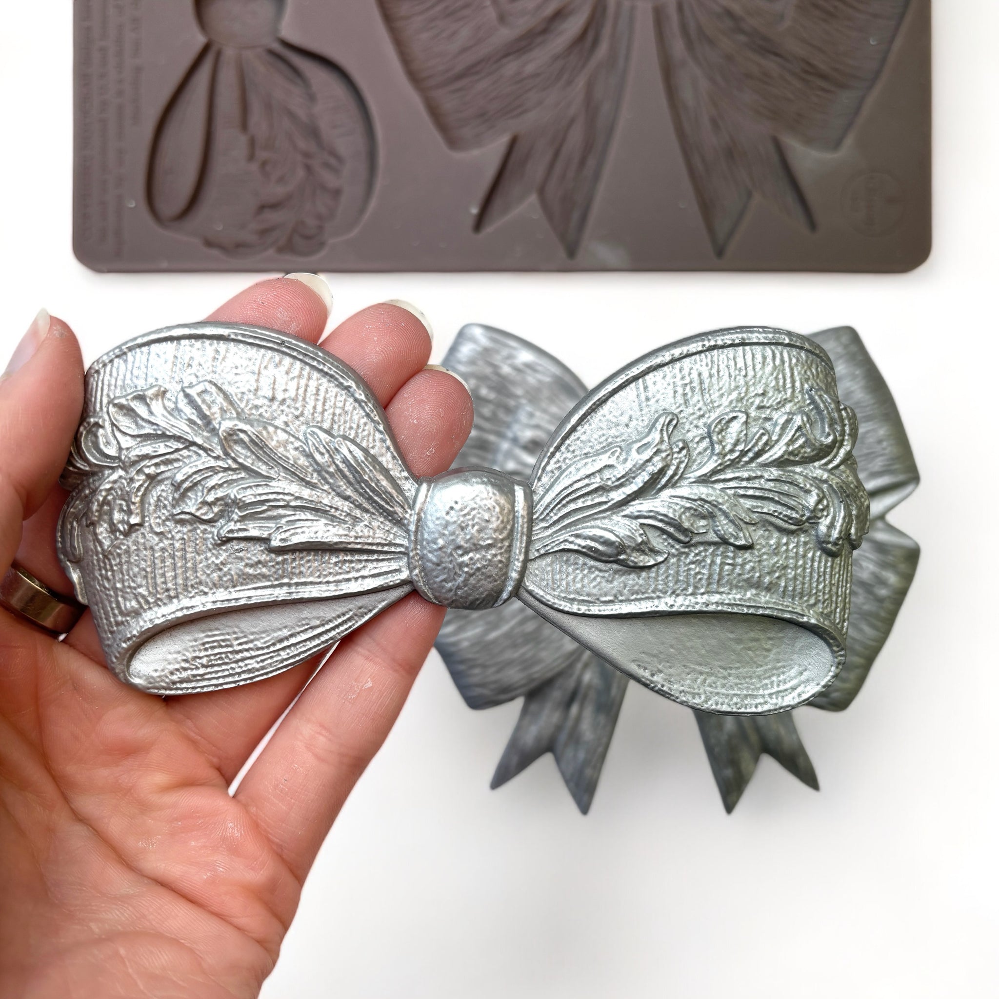 A close-up of a hand holding a silver colored casting of a bow created from ReDesign with Prima's Vintage Opulence silicone mold is against a white background.