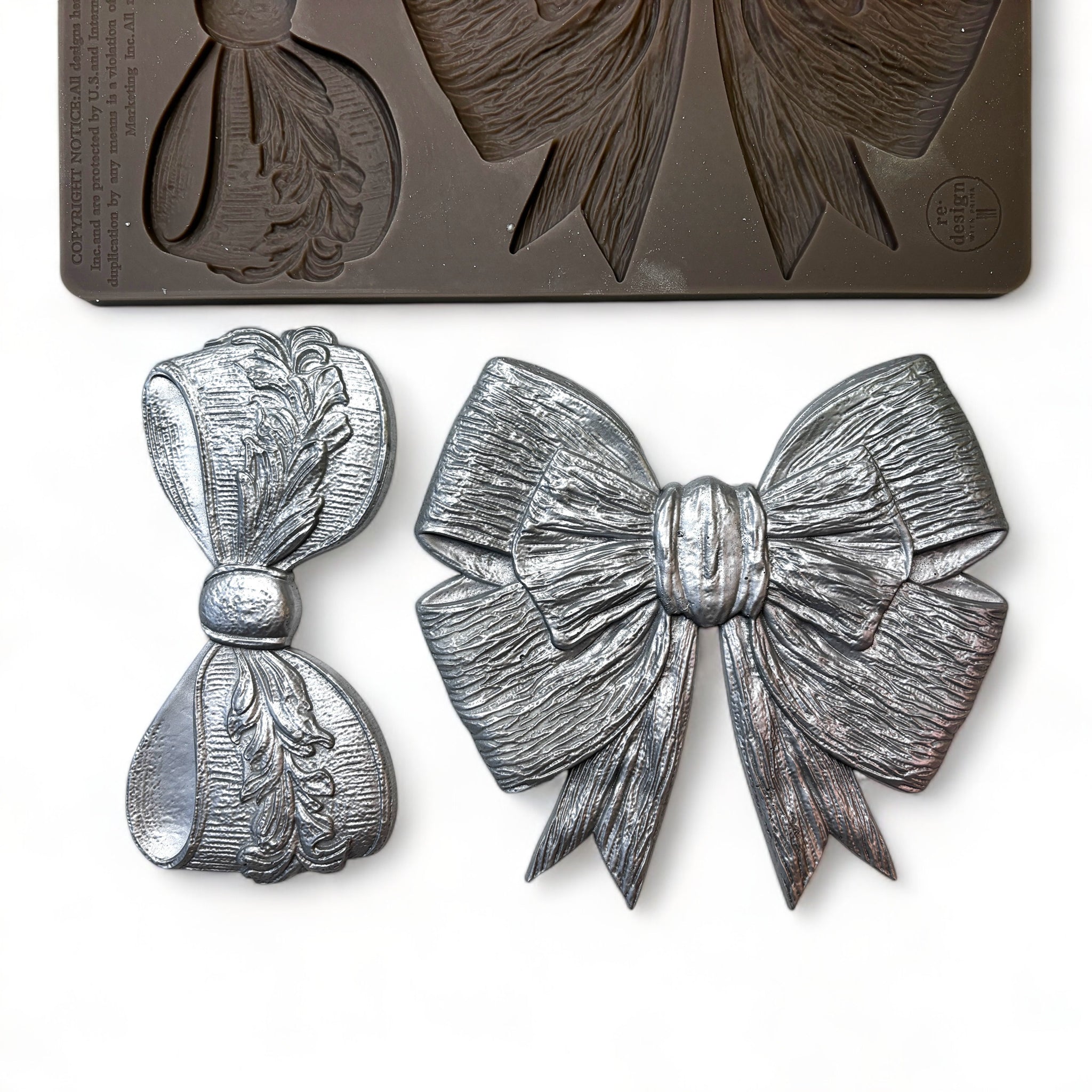 A brown silicone mold and silver colored castings of 2 different style ribbon bows are against a white background.