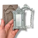 A silicone mold and silver colored castings featuring an intricate frame and smaller solid mirror-like frame are against a white background. A hand is shown holding the mirror-like frame.