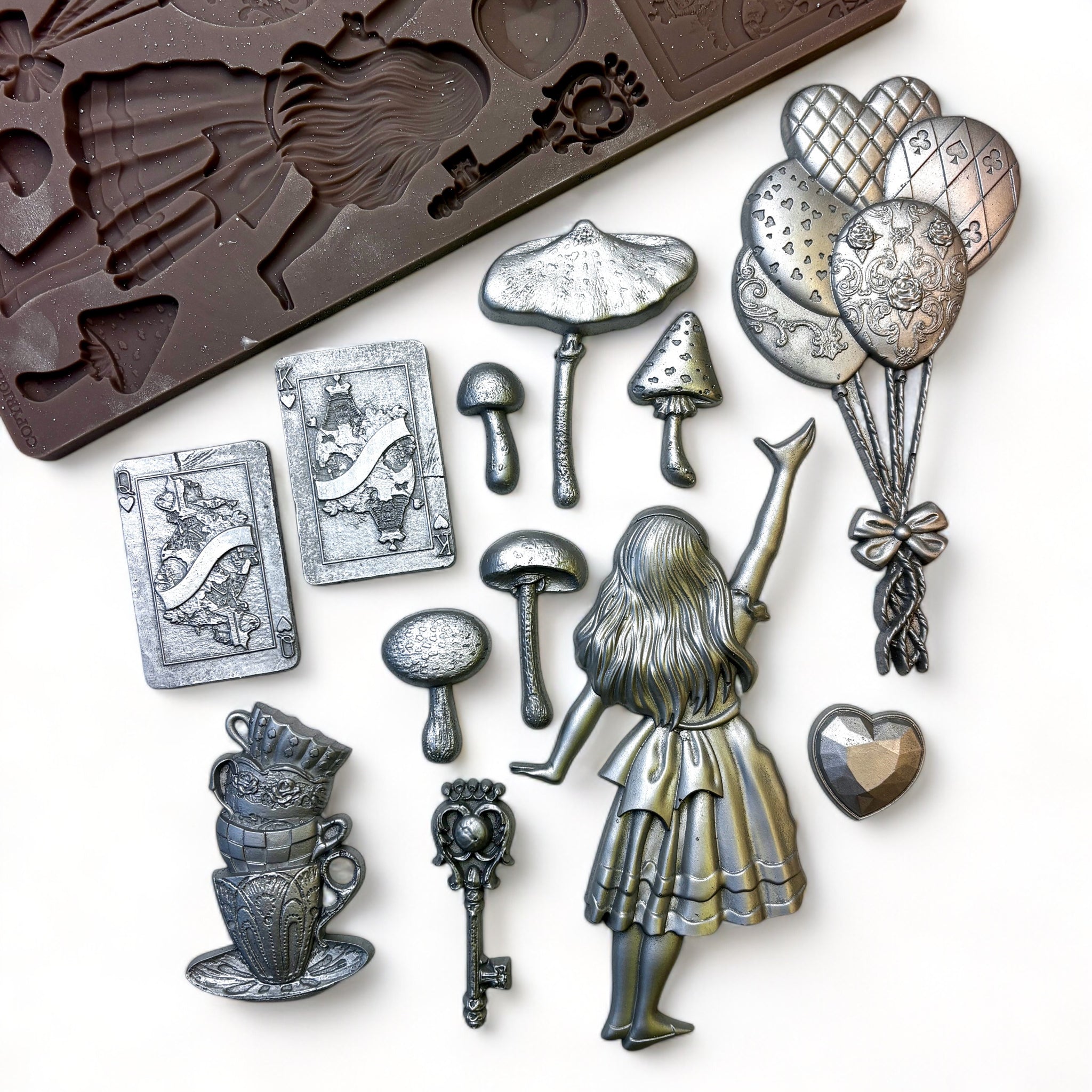 A brown silicone mold and silvered colored silicone castings featuring Alice in Wonderland, a bouquet of balloons, stack of teacups, mushrooms, playing cards, and a heart are against a white background.