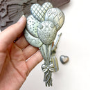 A close-up of a hand holding a silver colored casting of a bouquet of balloons created from ReDesign with Prima's Whimsy Craft Chronicles silicone mold is against a white background.