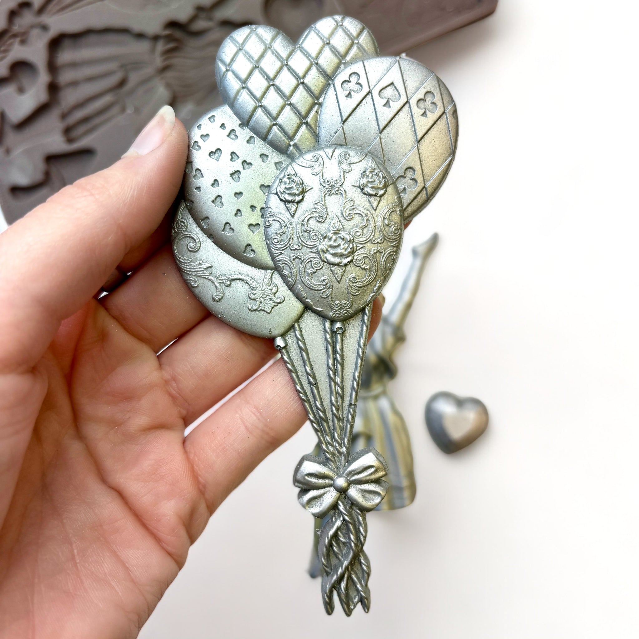 A close-up of a hand holding a silver colored casting of a bouquet of balloons created from ReDesign with Prima's Whimsy Craft Chronicles silicone mold is against a white background.