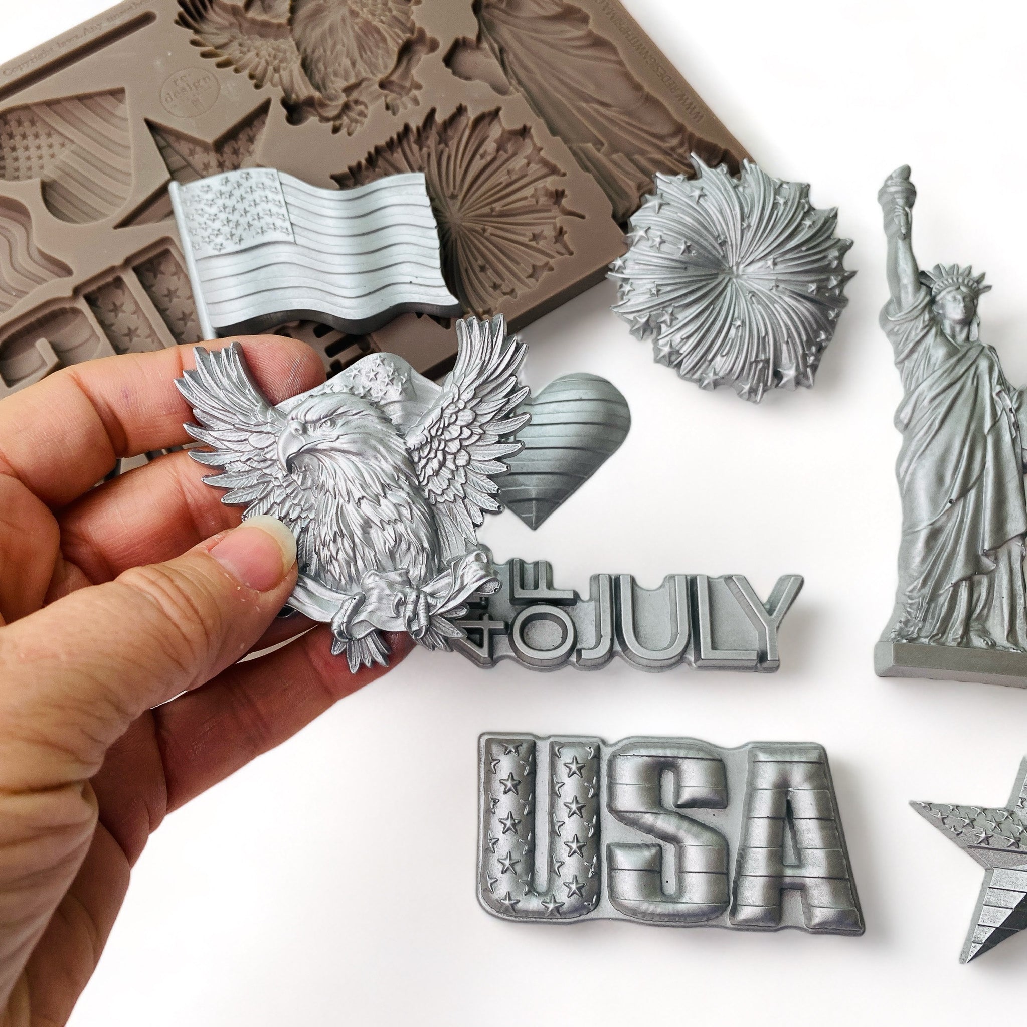 A brown silicone mold and silver colored castings of ReDesign with Prima's Happy 4th silicone mold are against a white background. A hand is shown holding the casting of the bald eagle design from the mold.