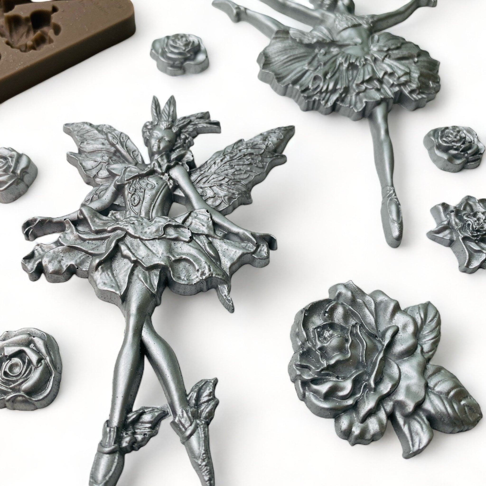 A close-up of a brown silicone mold and silver colored castings of 2 dancing ballerinas, a fairy ballerina, and 9 varying sizes of roses are against a white background.