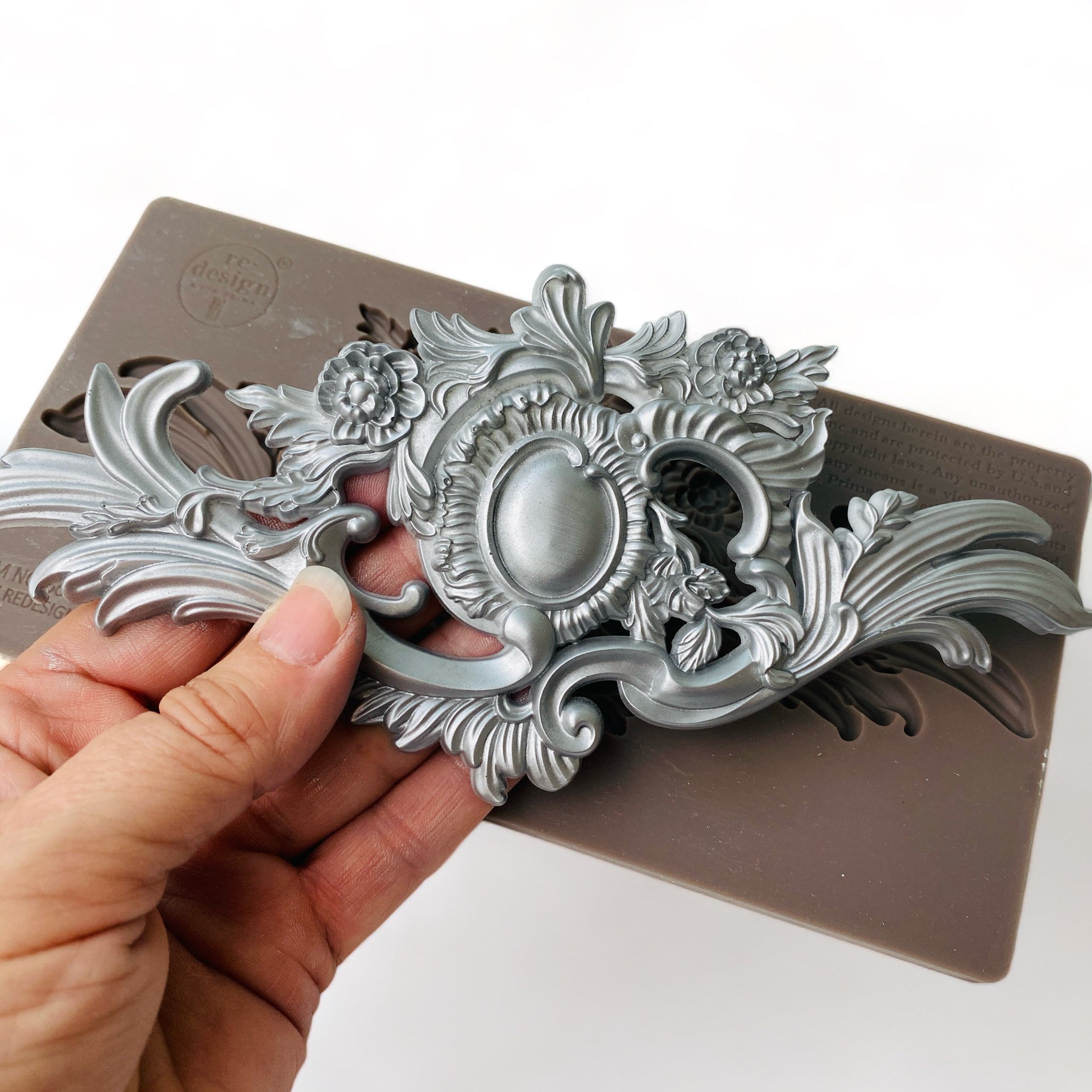 A brown silicone mold and a hand holding a silver colored casting that features a unique ornate flourish and central medallion are against a white background.