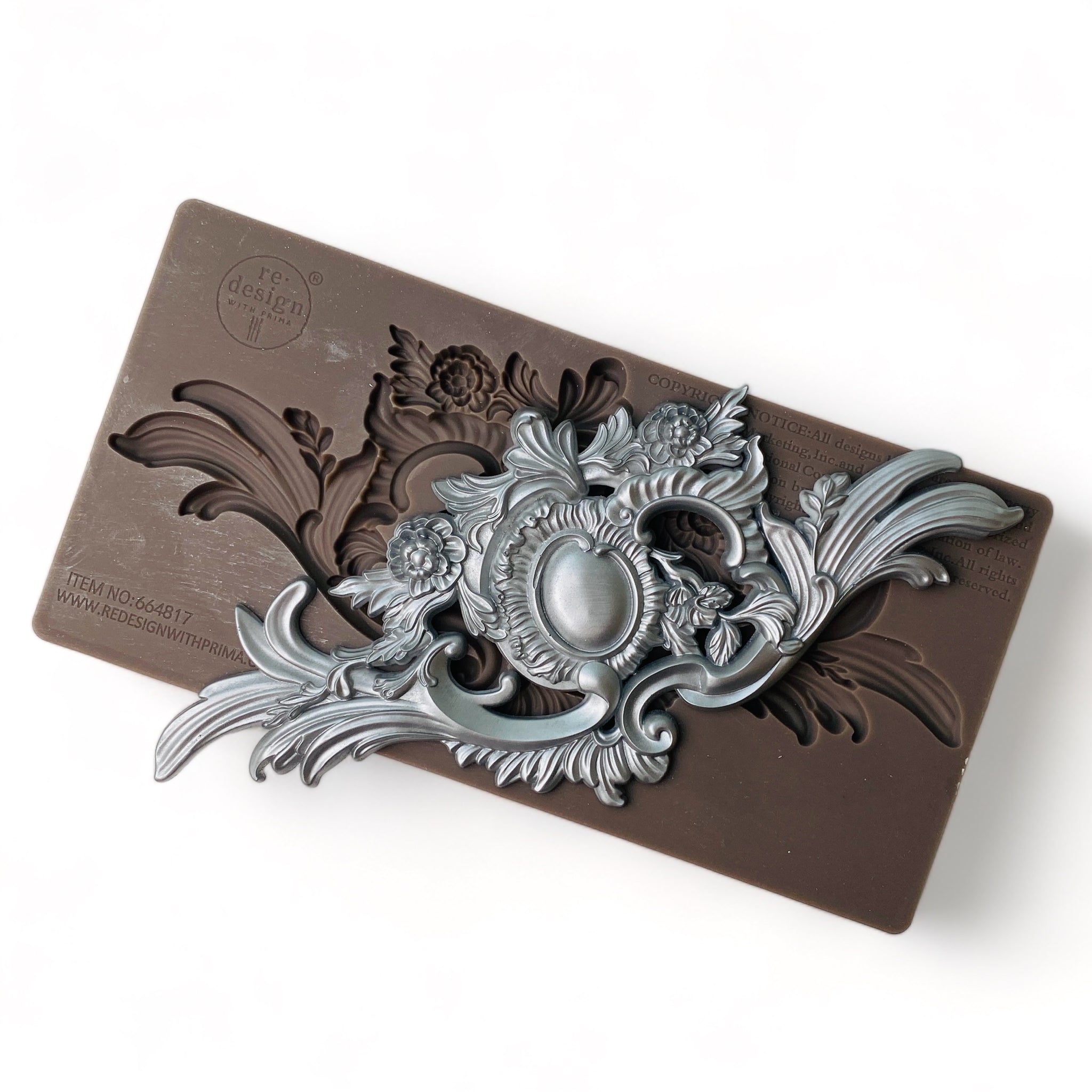 A brown silicone mold and silver colored casting that features a unique ornate flourish and central medallion are against a white background.
