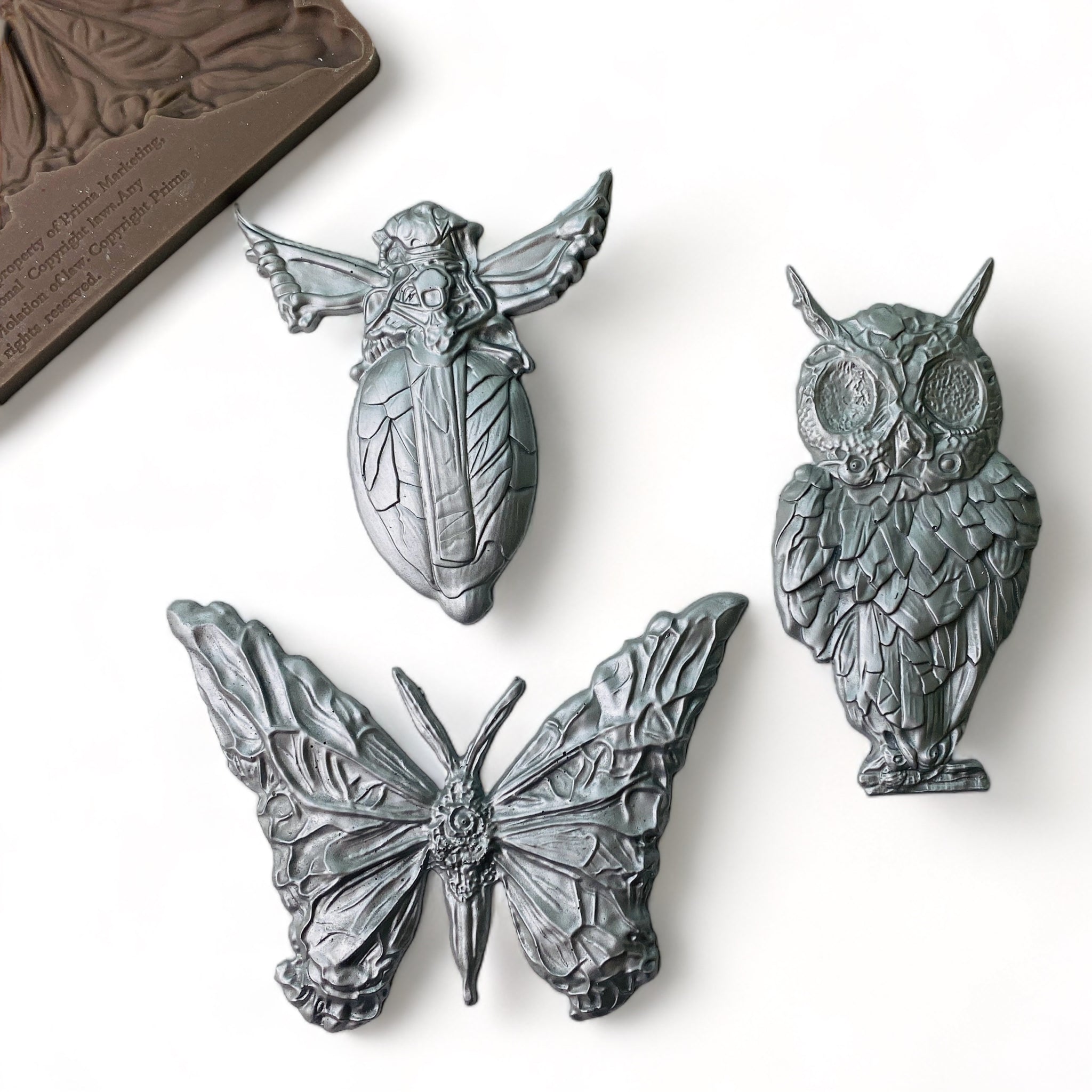 A brown silicone mold and silver colored castings of a butterfly, owl, and flying beetle are against a white background.