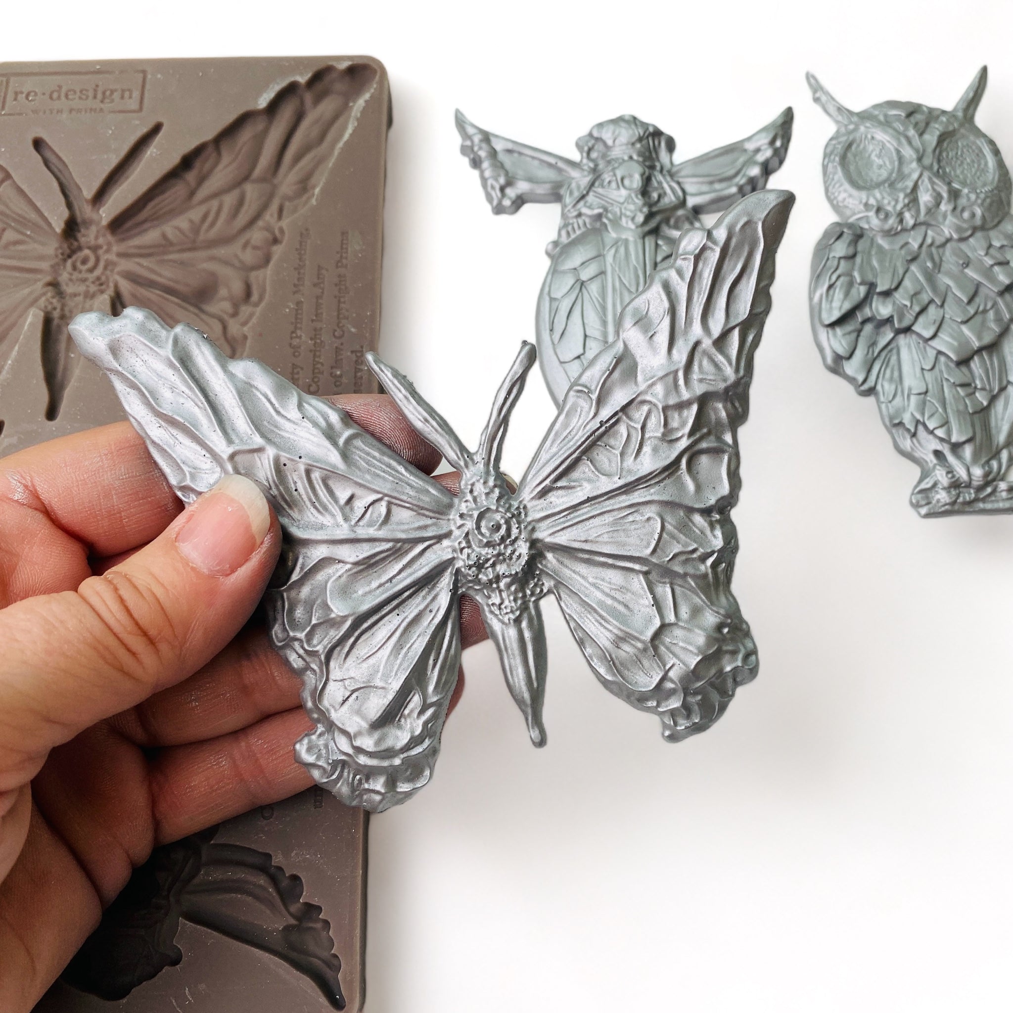 A brown silicone mold and silver colored castings of a butterfly, owl, and flying beetle are against a white background. A hand is shown holding the moth casting.