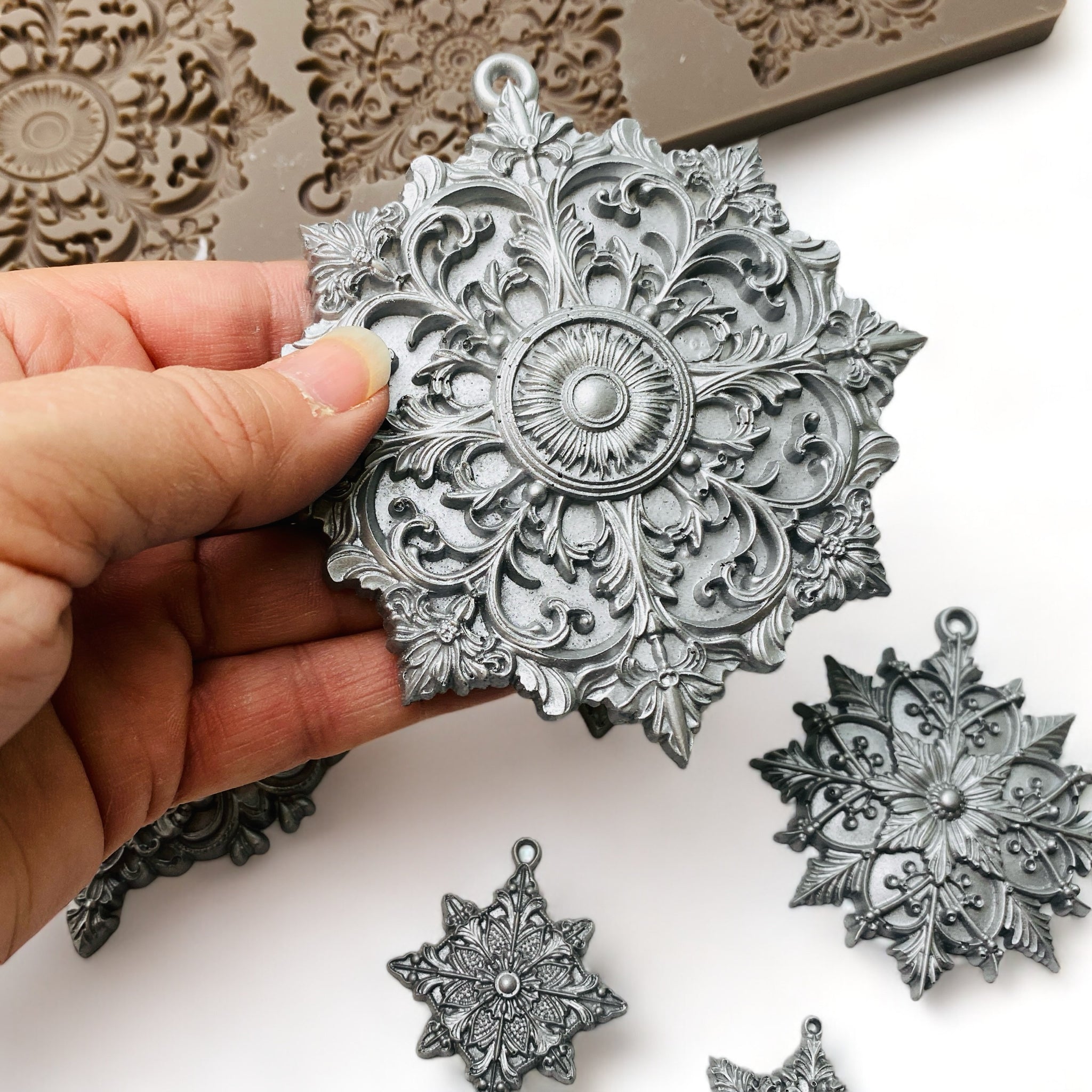 A hand is shown holding a silver colored casting of a large snowflake from ReDesign with Prima's Dazzling Ornates silicone mold.