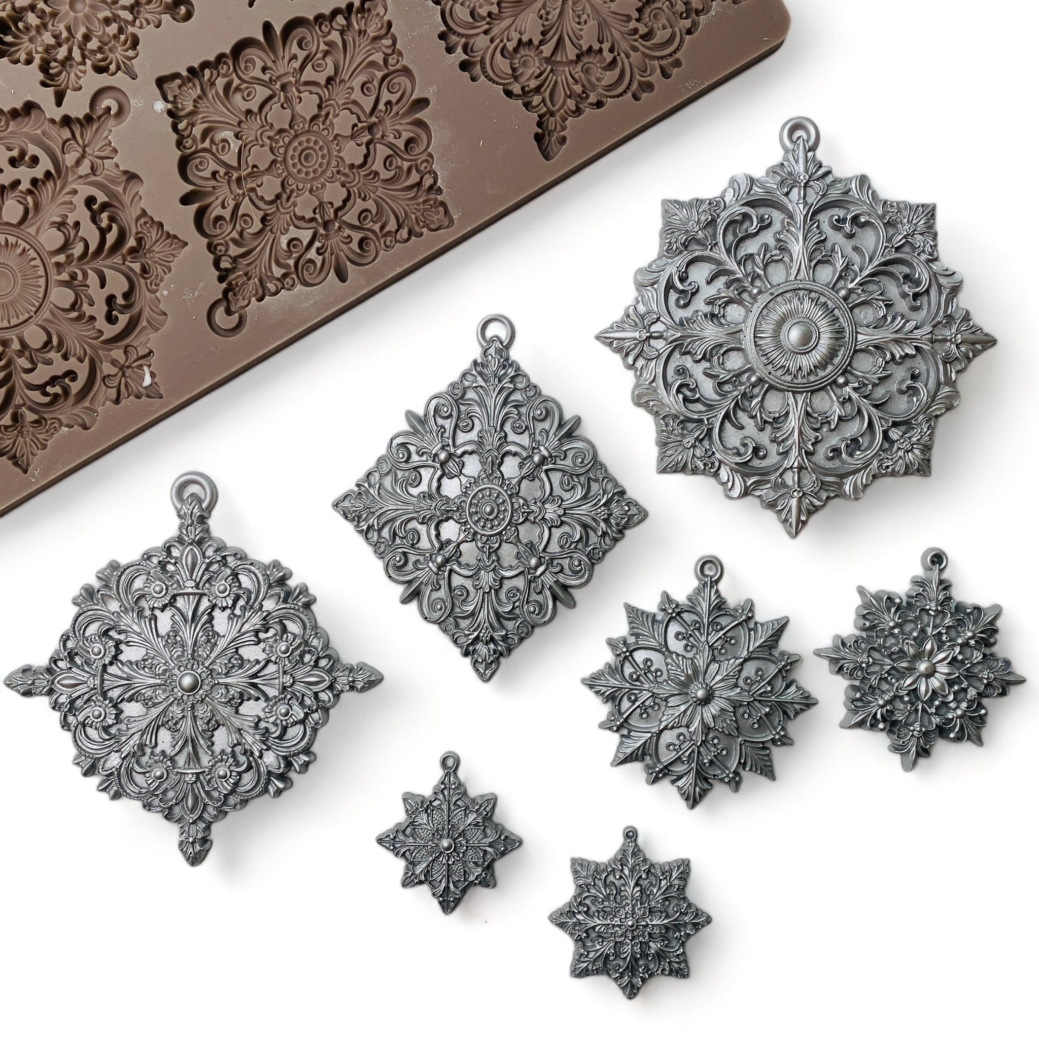 A brown silicone mold and silver colored castings of 3 large and 4 smaller ornately decorated snowflakes are against a white background.