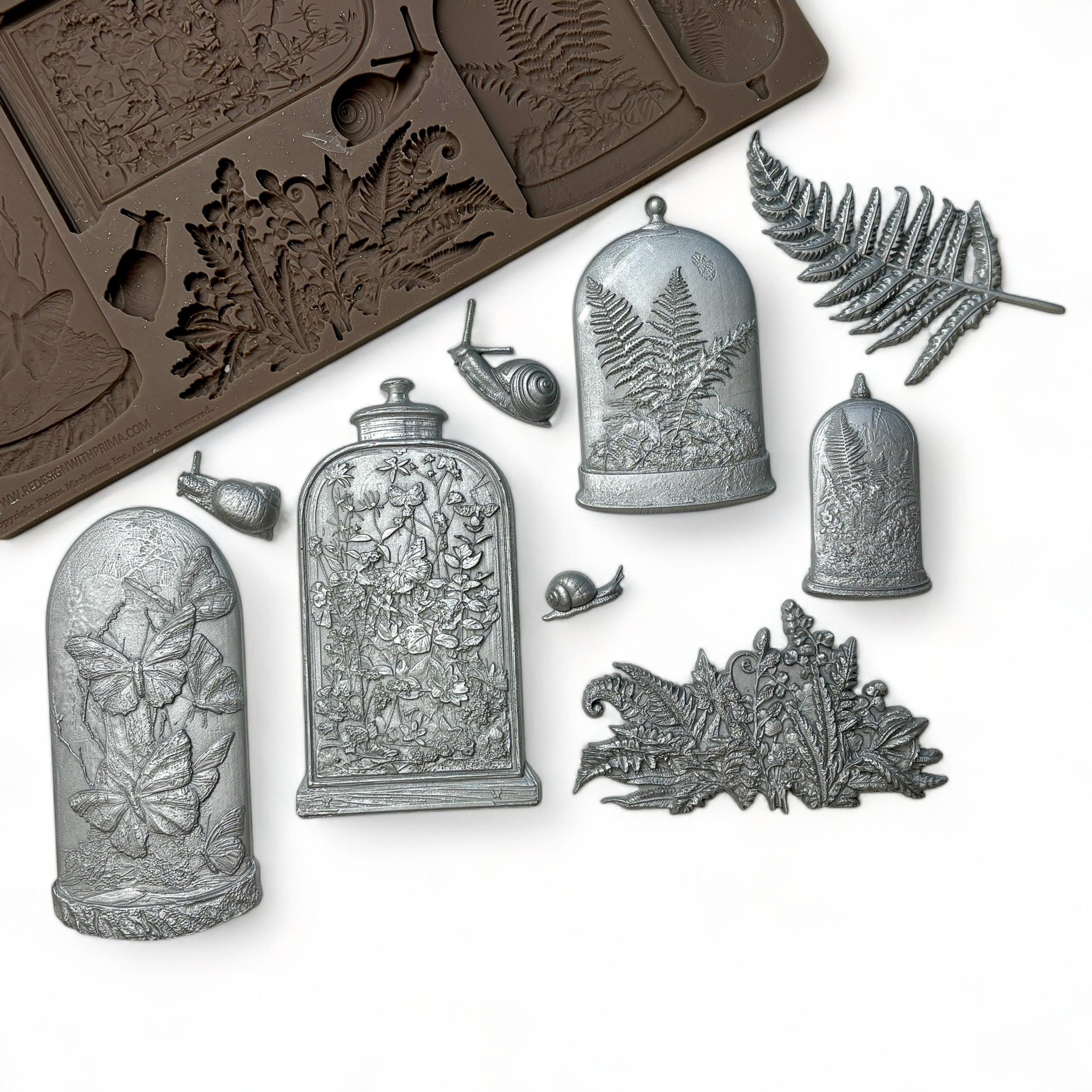 A brown silicone mold and silver colored castings of 4 Victorian cloches with plants and butterflies in them, and ferns and snails are against a white background.