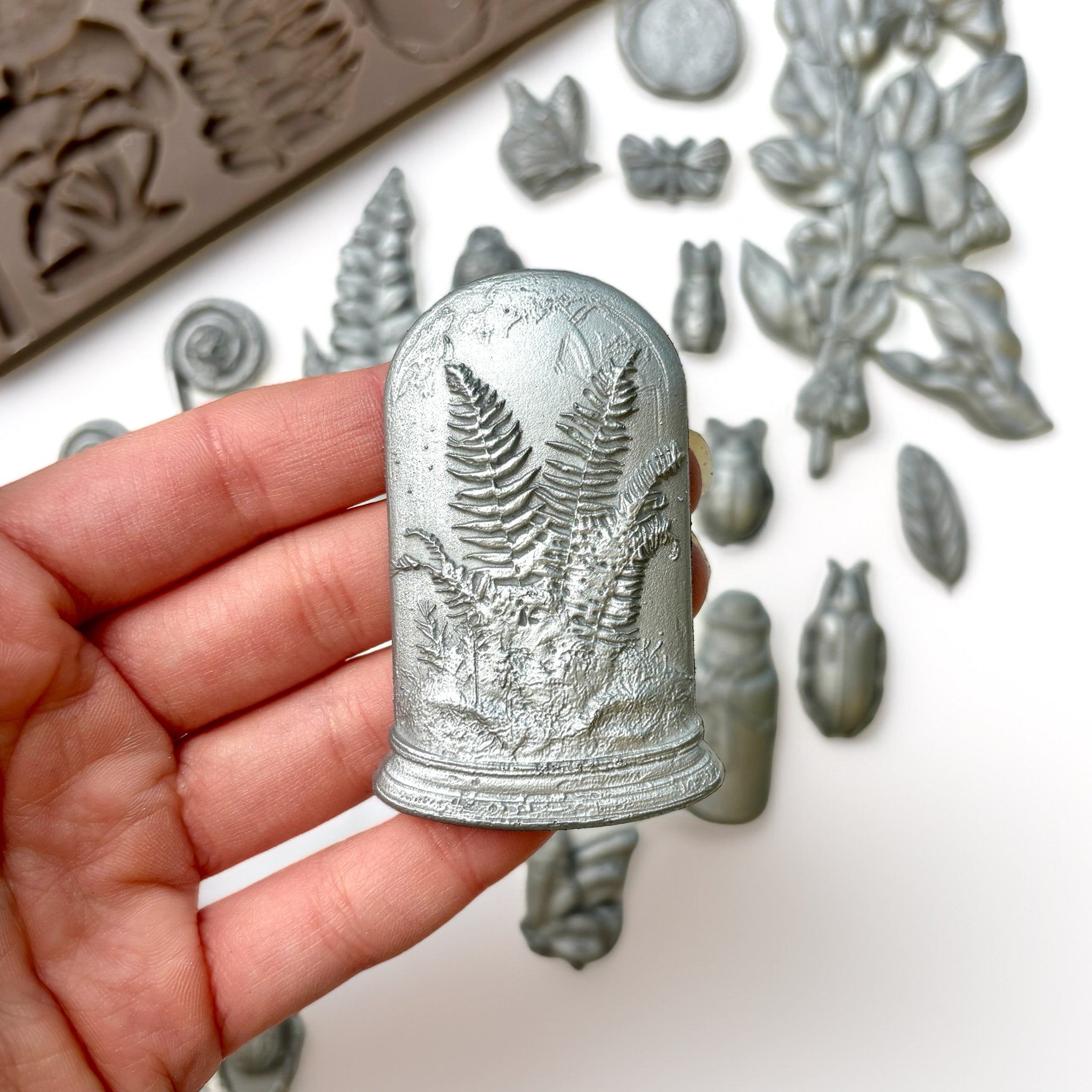 A brown silicone mold and silver colored castings of 4 Victorian cloches with plants and butterflies in them, and ferns and snails are against a white background. A hand is shown holding one of the cloches.