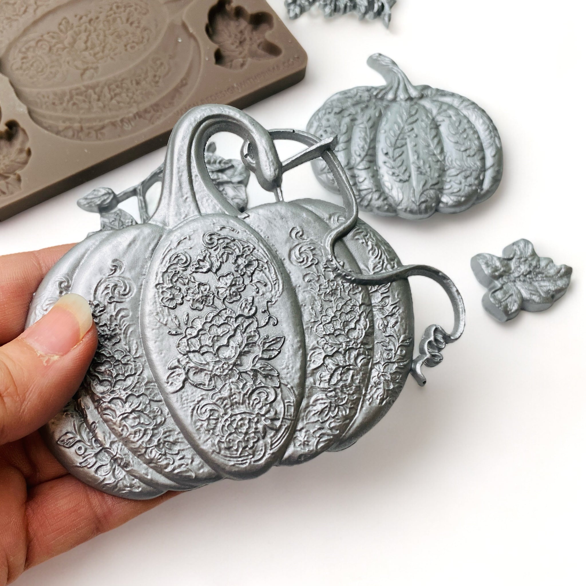 A hand is shown holding a silver colored casting of a pumpkin from ReDesign with Prima's Falling for Fall silicone mold.