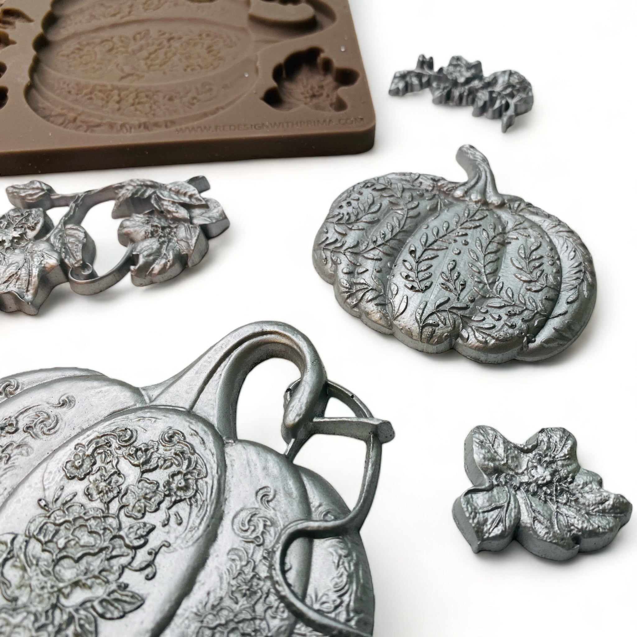 A close-up of a brown silicone mold and silver colored castings of 3 pumpkins and some loose vines and leaves are against a white background.