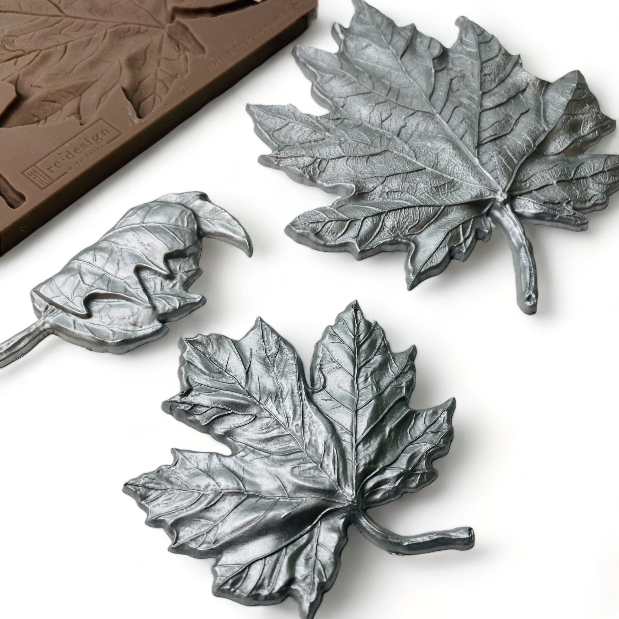 A brown silicone mold and silver colored castings of 3 varying size maple leaves are against a white background.