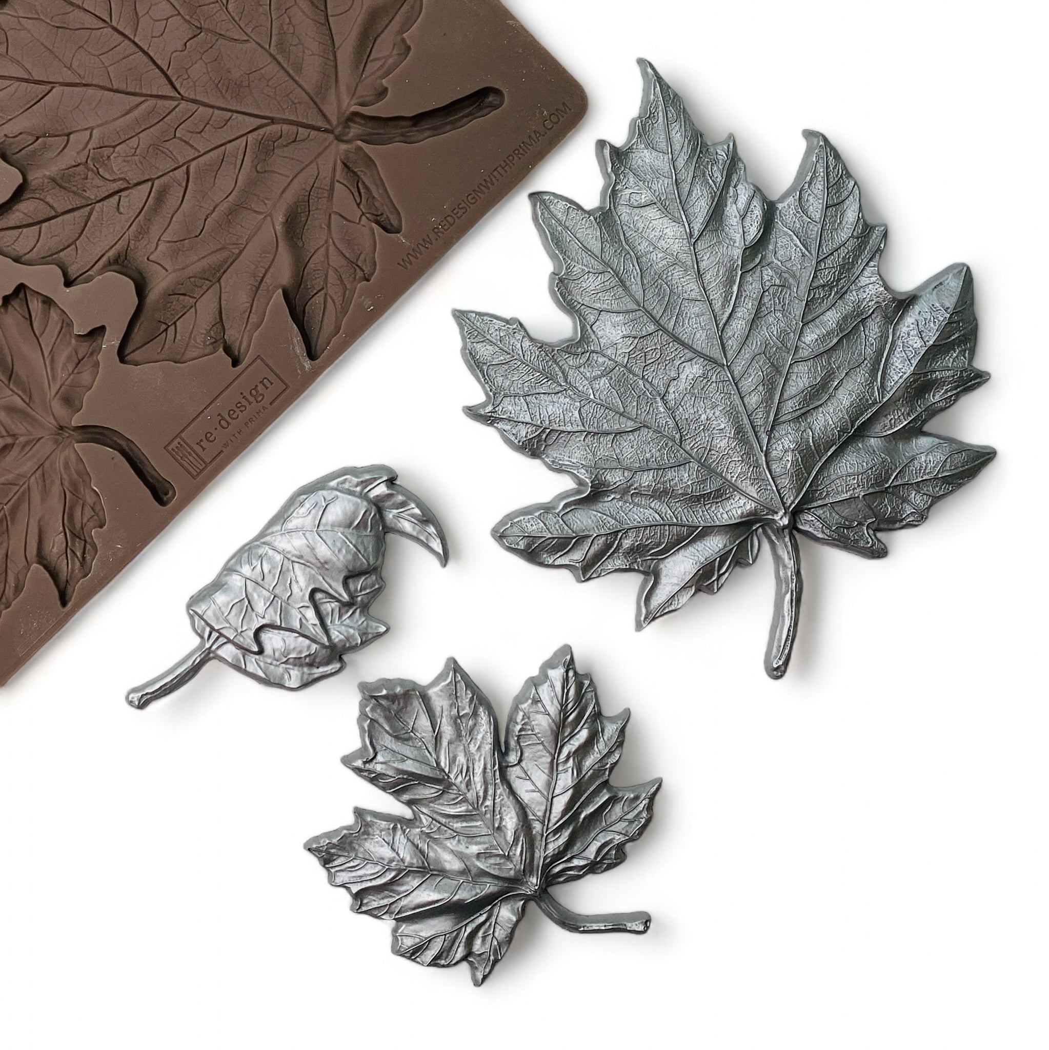 A brown silicone mold and silver colored castings of 3 varying size maple leaves are against a white background.