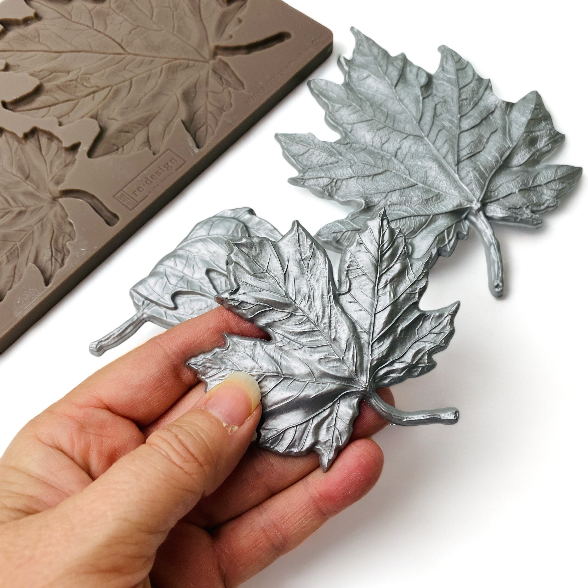 A hand is shown holding a silver colored casting of a leaf from ReDesign with Prima's Falling Leaves Fantasy silicone mold.