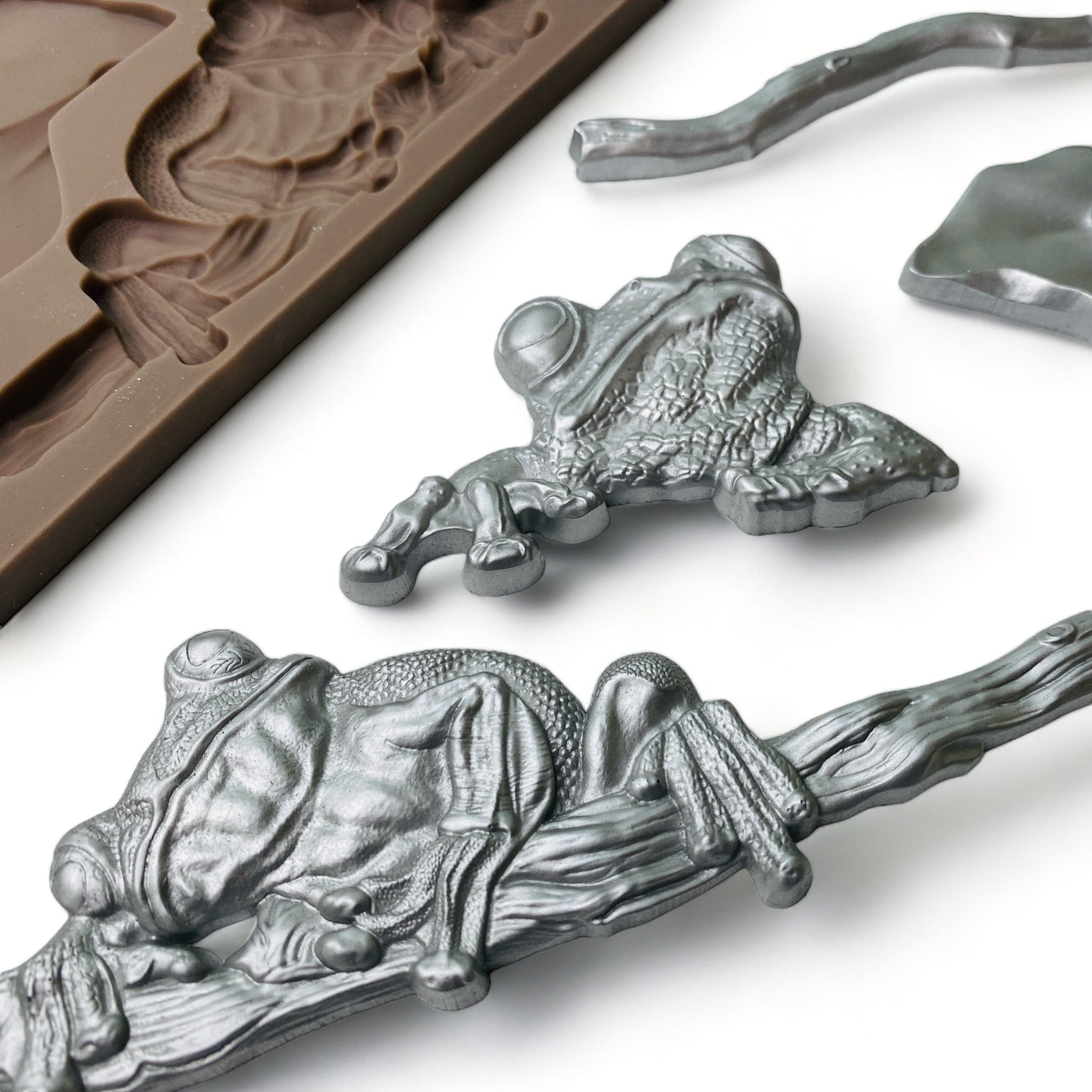 Close-up of a brown silicone mold and silver colored castings of 3 frogs; 1 on a branch, 1 on a leaf, and 1 separate with a loose branch are against a white background.