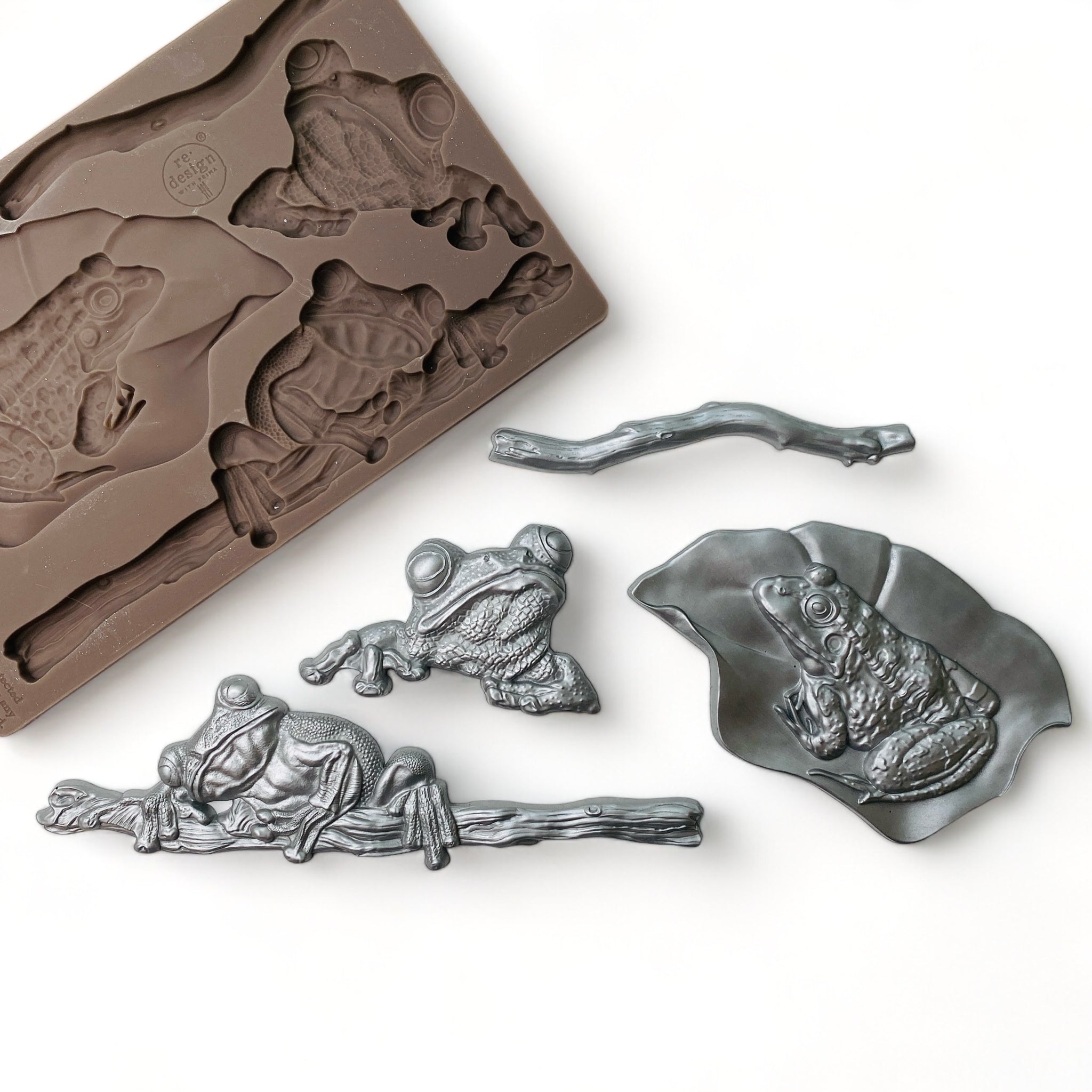 A brown silicone mold and silver colored castings of 3 frogs; 1 on a branch, 1 on a leaf, and 1 separate with a loose branch are against a white background.