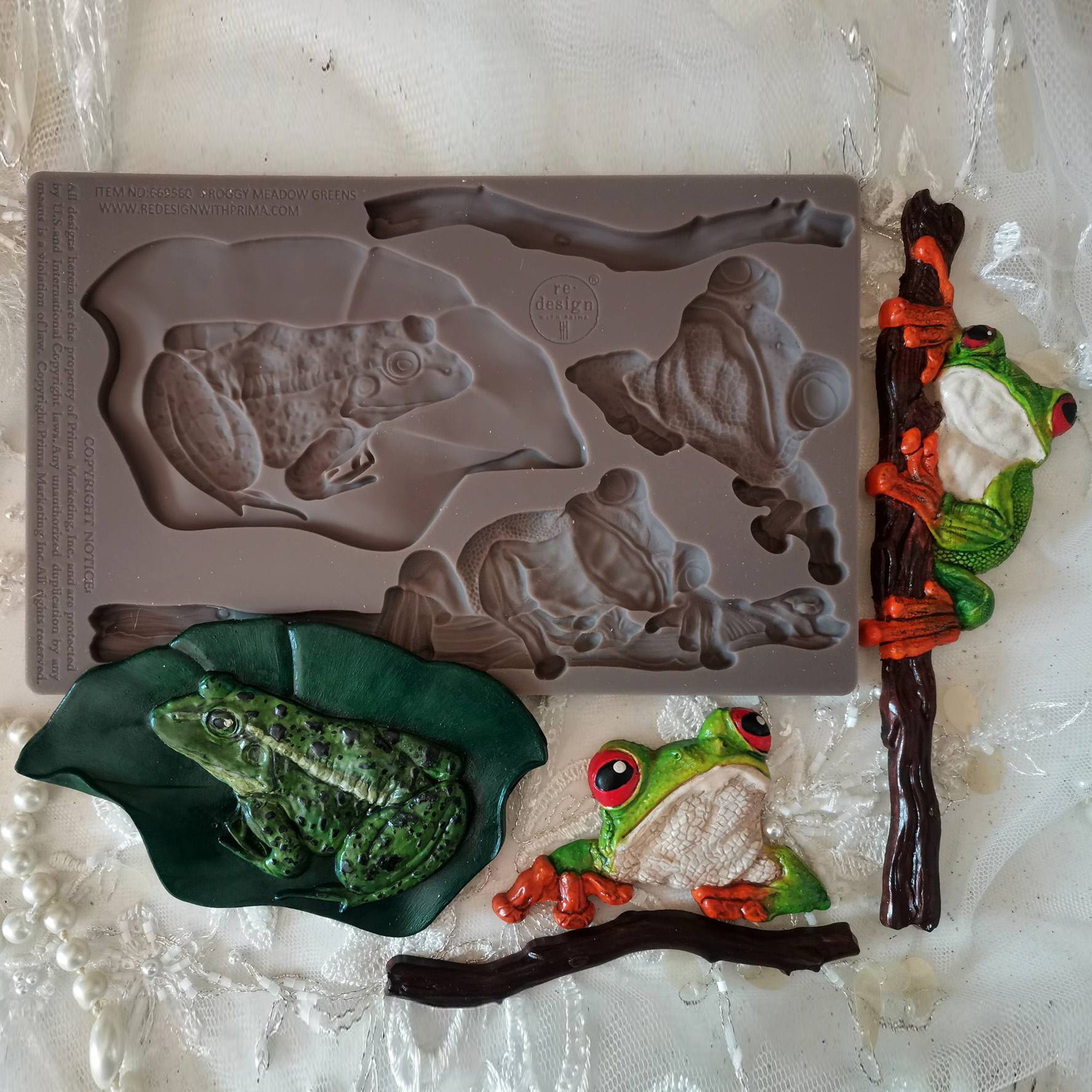 A brown silicone mold and colorful castings of 3 frogs on branches and leaves are against a white background.