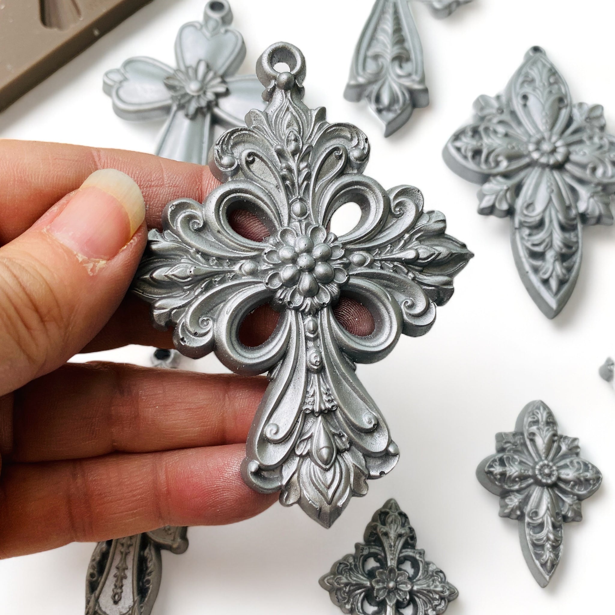 A hand is shown holding a silver colored casting of an ornate cross from ReDesign with Prima's Holy Harmony silicone mold.