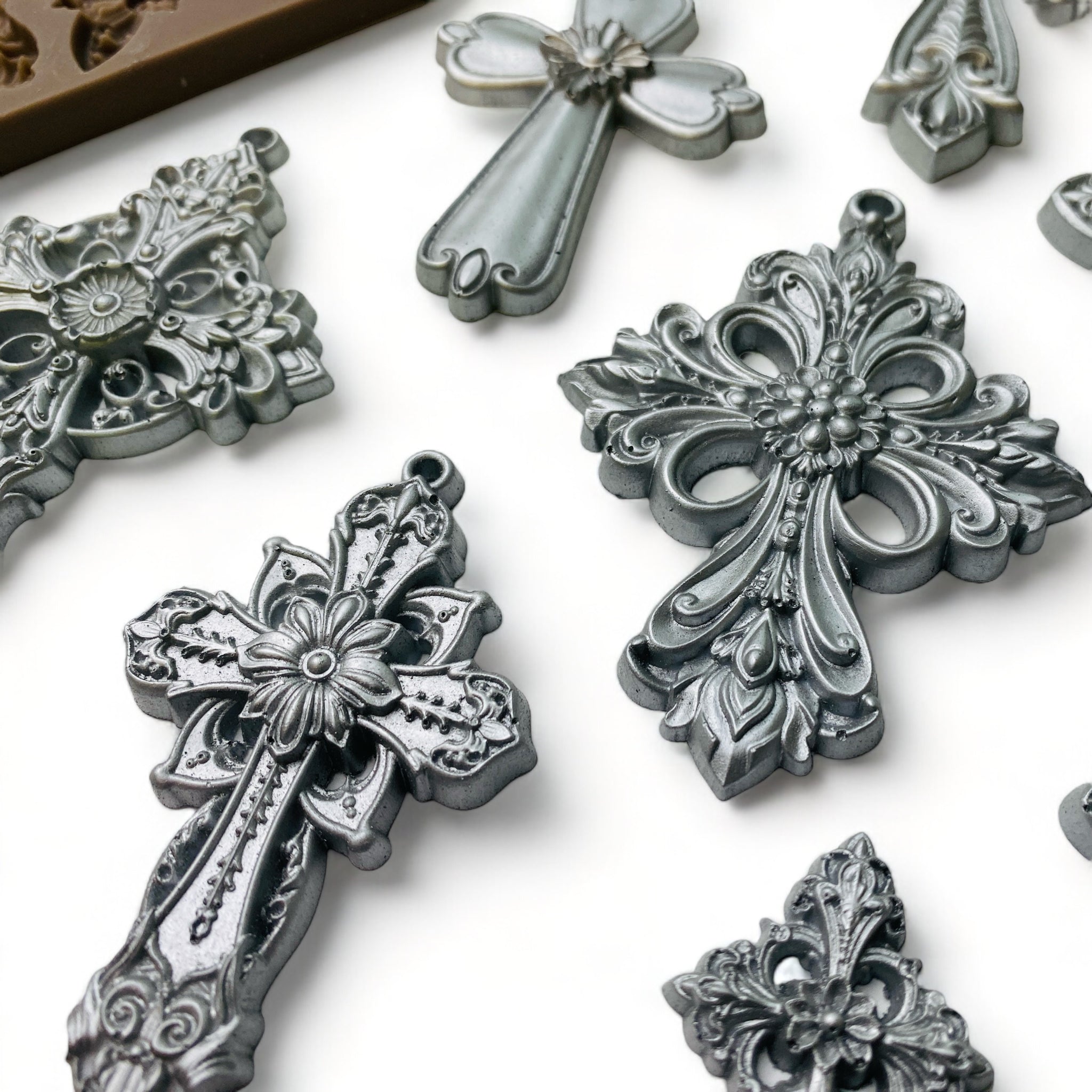 Close-up of a brown silicone mold and silver colored castings of 9 varying sizes of ornate vintage crosses are against a white background.