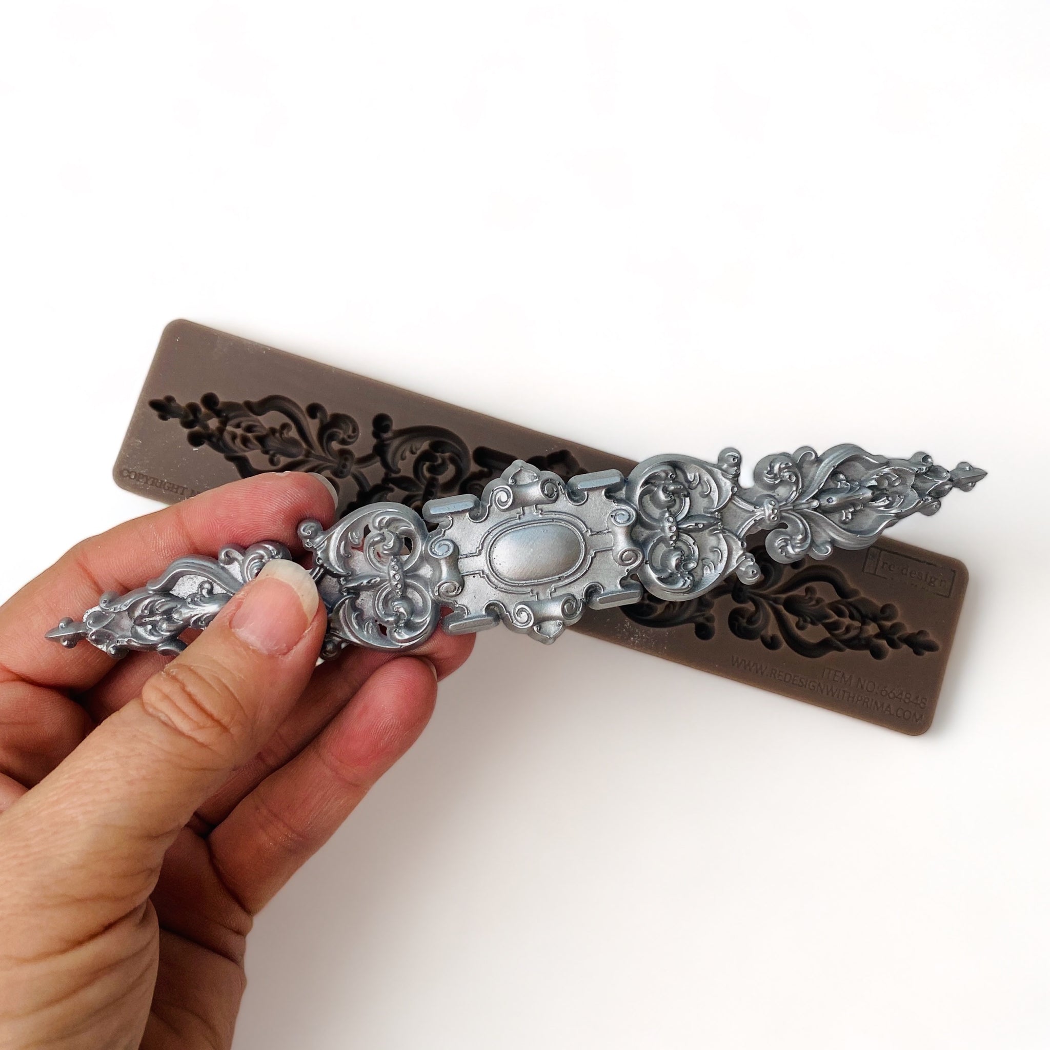 A brown silicone mold and a hand holding a silver colored casting of a long, ornate, scroll decorative center medallion are against a white background.